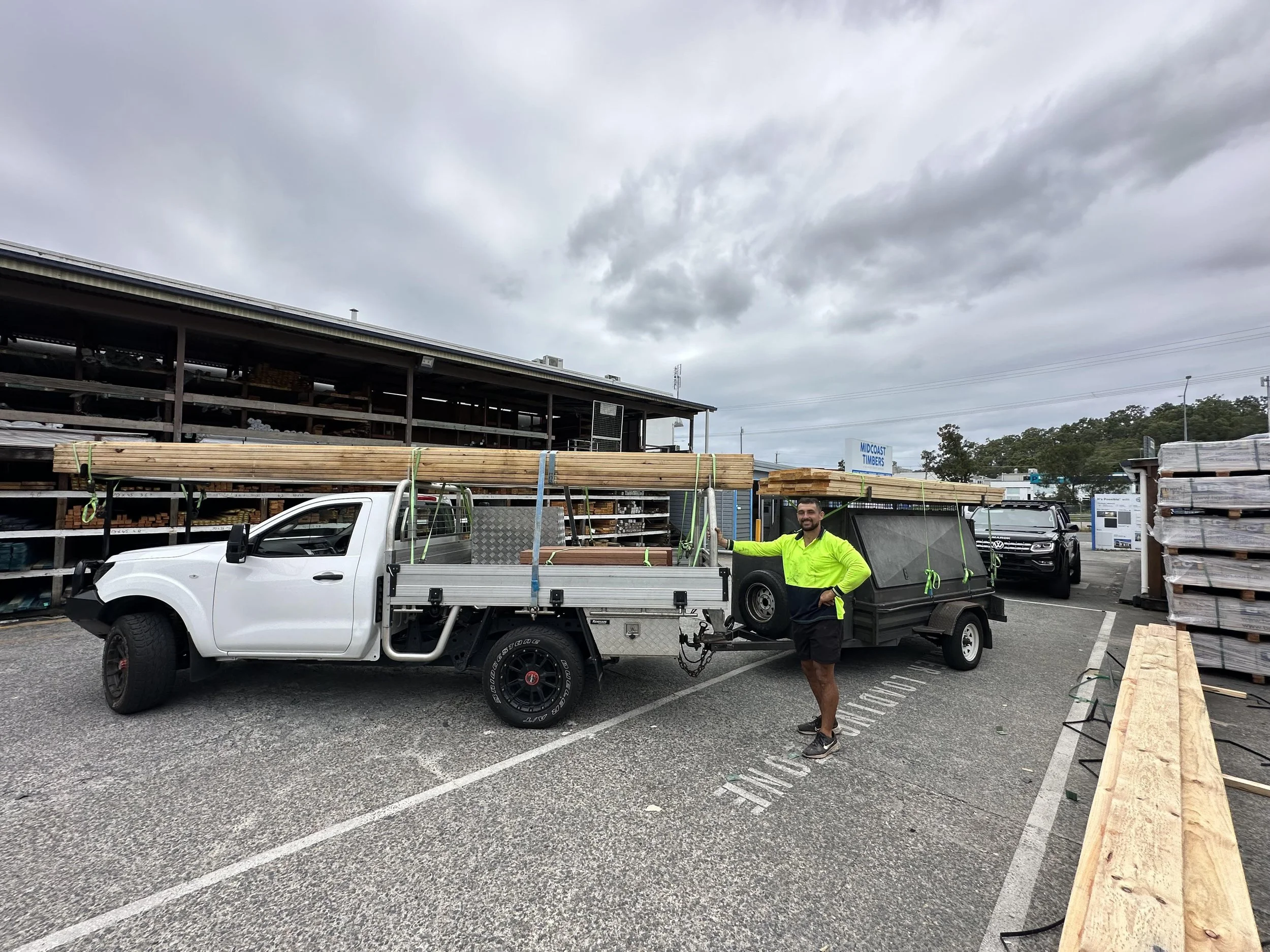 A man in a neon yellow jacket standing next to a white pickup truck with a flatbed, loaded with long wooden planks, parked in a hardware store lot. There are stacks of wood and other building materials around him, with overcast skies above.