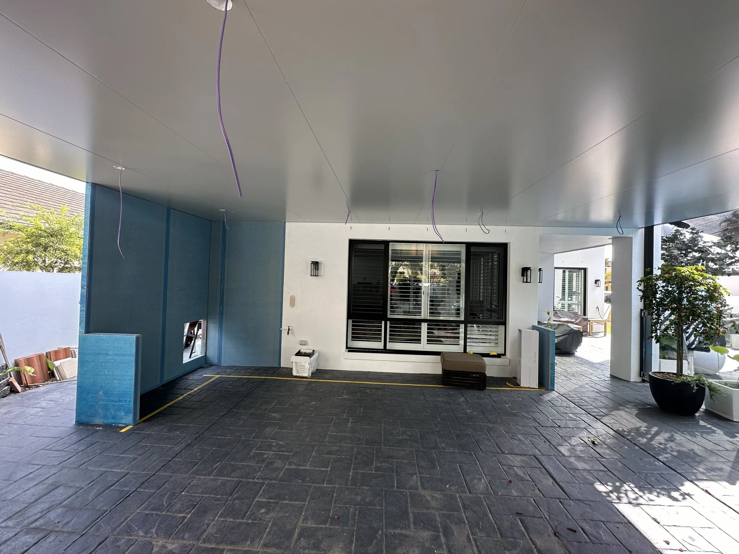 Construction site indoors with blue drywall, a large window with black framing, a bench, potted plants, and construction tools and materials scattered around.