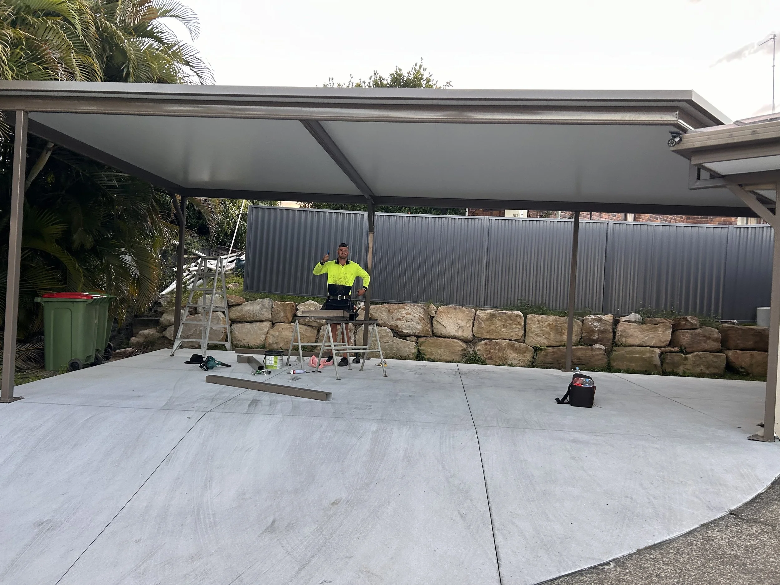 A man in a bright yellow safety shirt giving a thumbs up while working under a metal carport with tools and equipment on the ground.