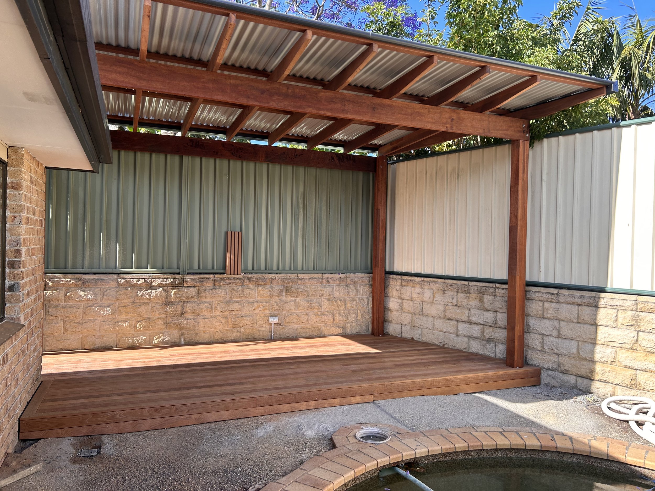 Backyard patio area with wooden deck and a covered pergola structure, surrounded by brick and stone walls, with some greenery and trees visible in the background.
