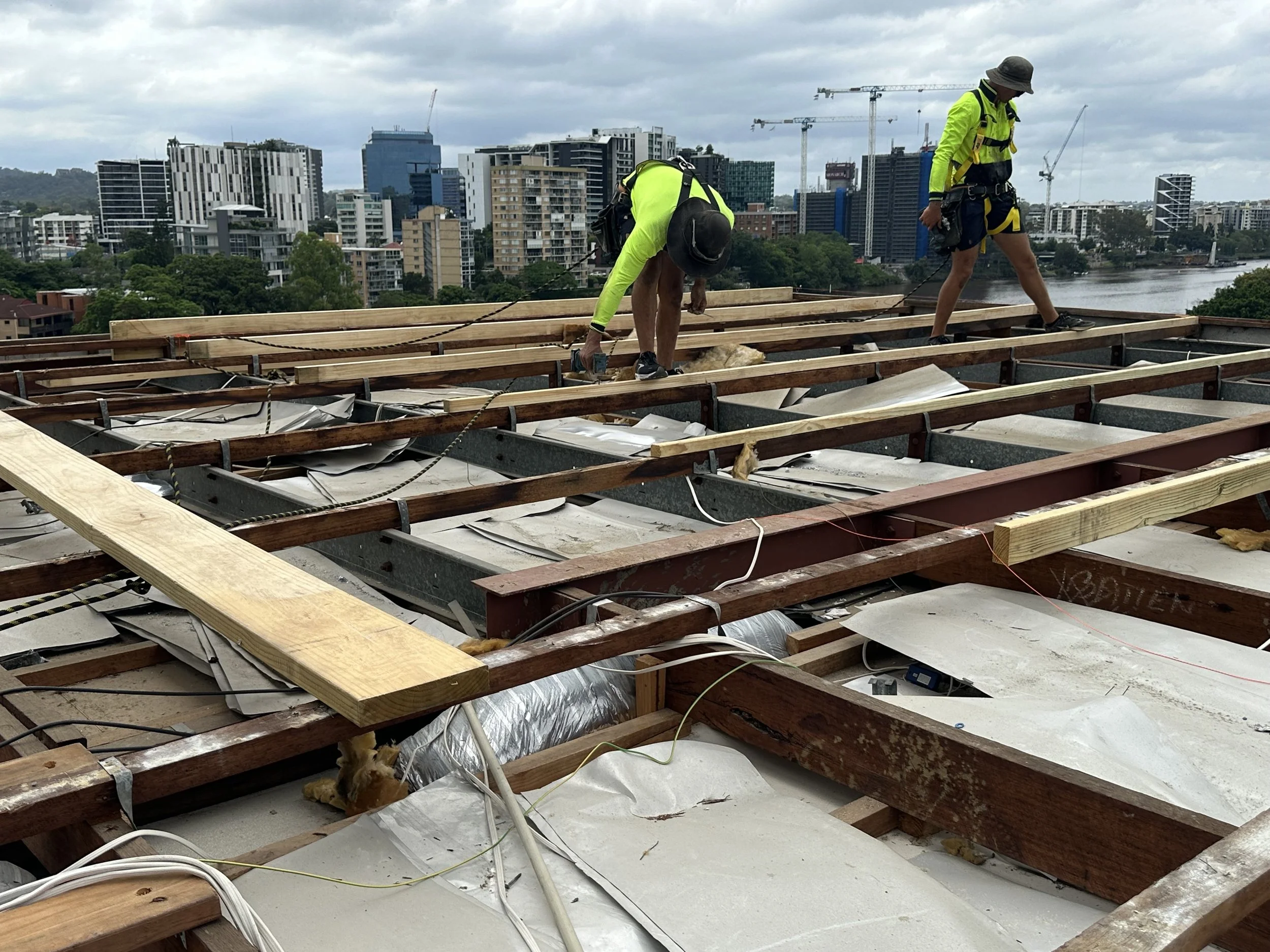 Two construction workers wearing high-visibility jackets, hats, and safety harnesses working on a building roof with the city skyline and river in the background.