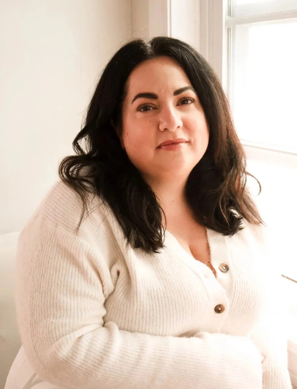 A woman with dark, wavy hair, wearing a cream-colored cardigan, sitting near a window with natural light.