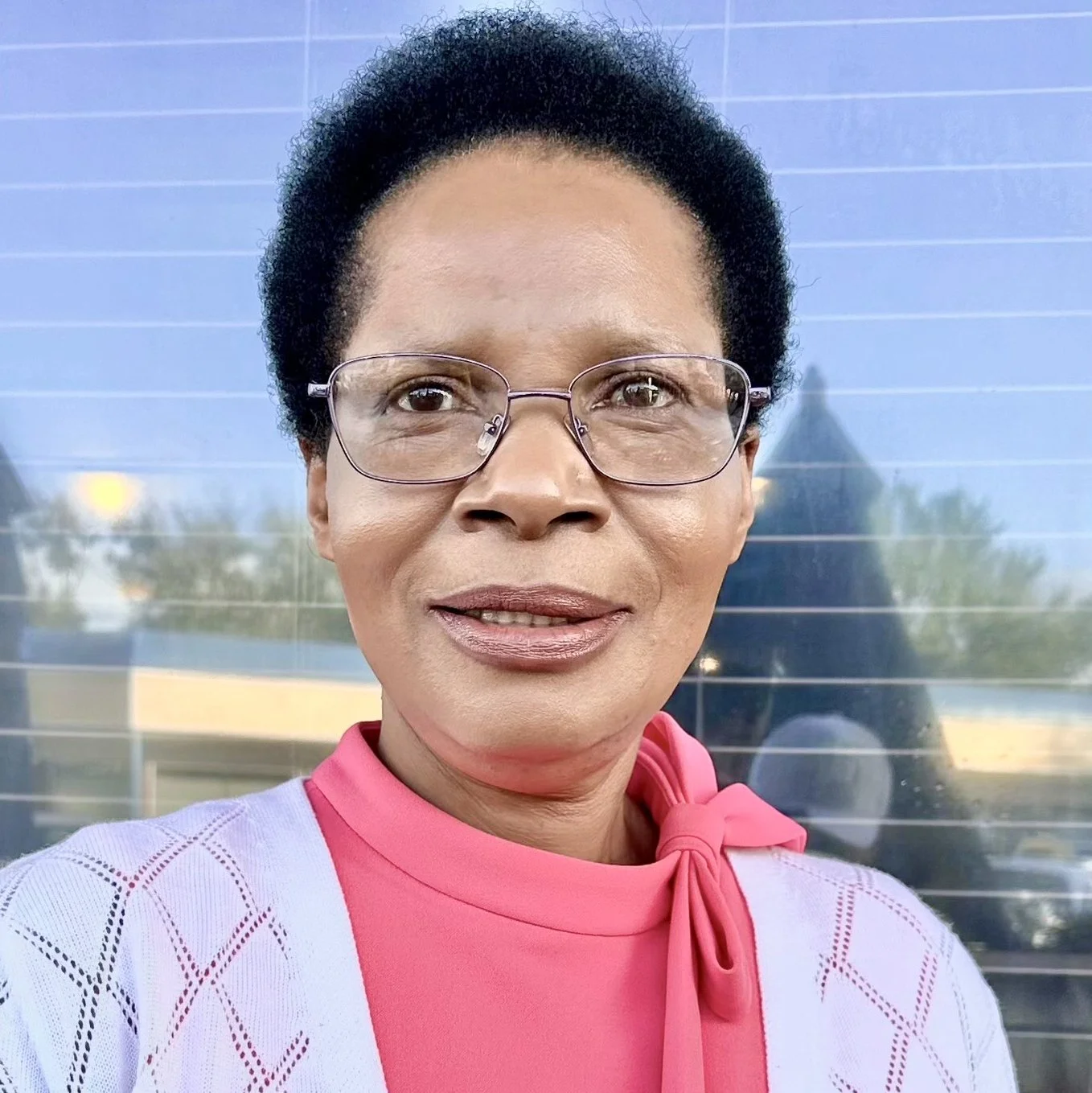 Close-up of a woman wearing glasses, a pink top with a bow at the neck, standing outdoors in front of a reflective glass background.
