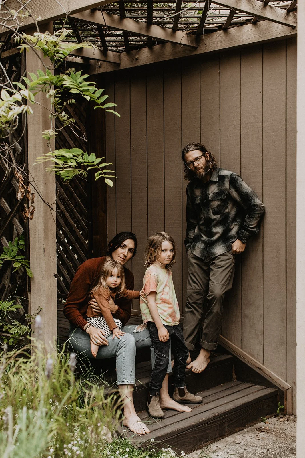 A family of four, consisting of a mother, father, and two young girls, poses outdoors on a small wooden deck with a brown wooden fence and a trellis overhead. The mother sits with one girl on her lap, the other girl stands nearby, and the father leans against the wall barefoot, wearing glasses and a plaid shirt.