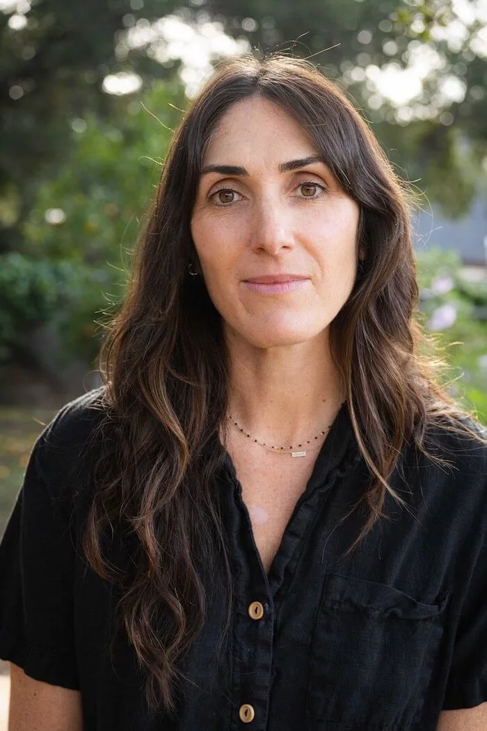A woman with long, wavy brown hair outdoors, wearing a black button-up shirt and a delicate necklace, with greenery in the background.