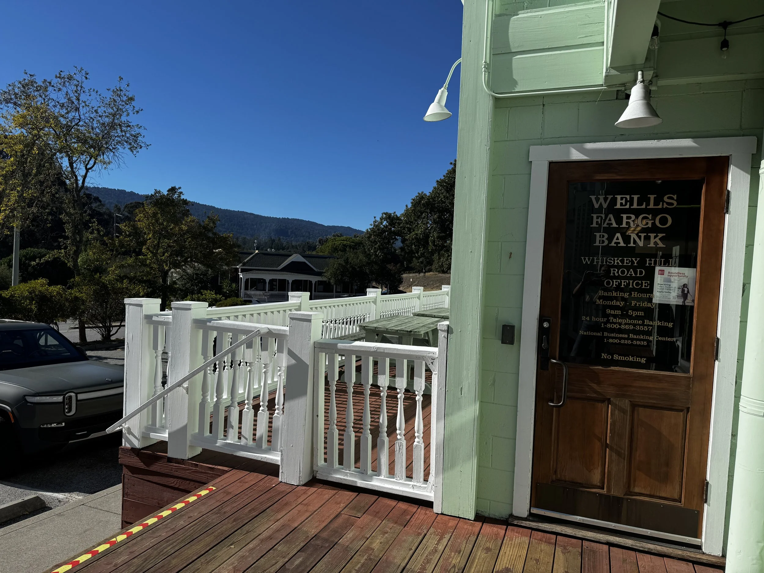 Exterior of a light green building with a wooden door labeled 'Wells Fargo Bank'. The building has white trim and outdoor lighting fixtures. In front, a white railing surrounds a small outdoor area with wooden decking. A car is parked nearby under a clear blue sky with some trees and distant mountains in the background.