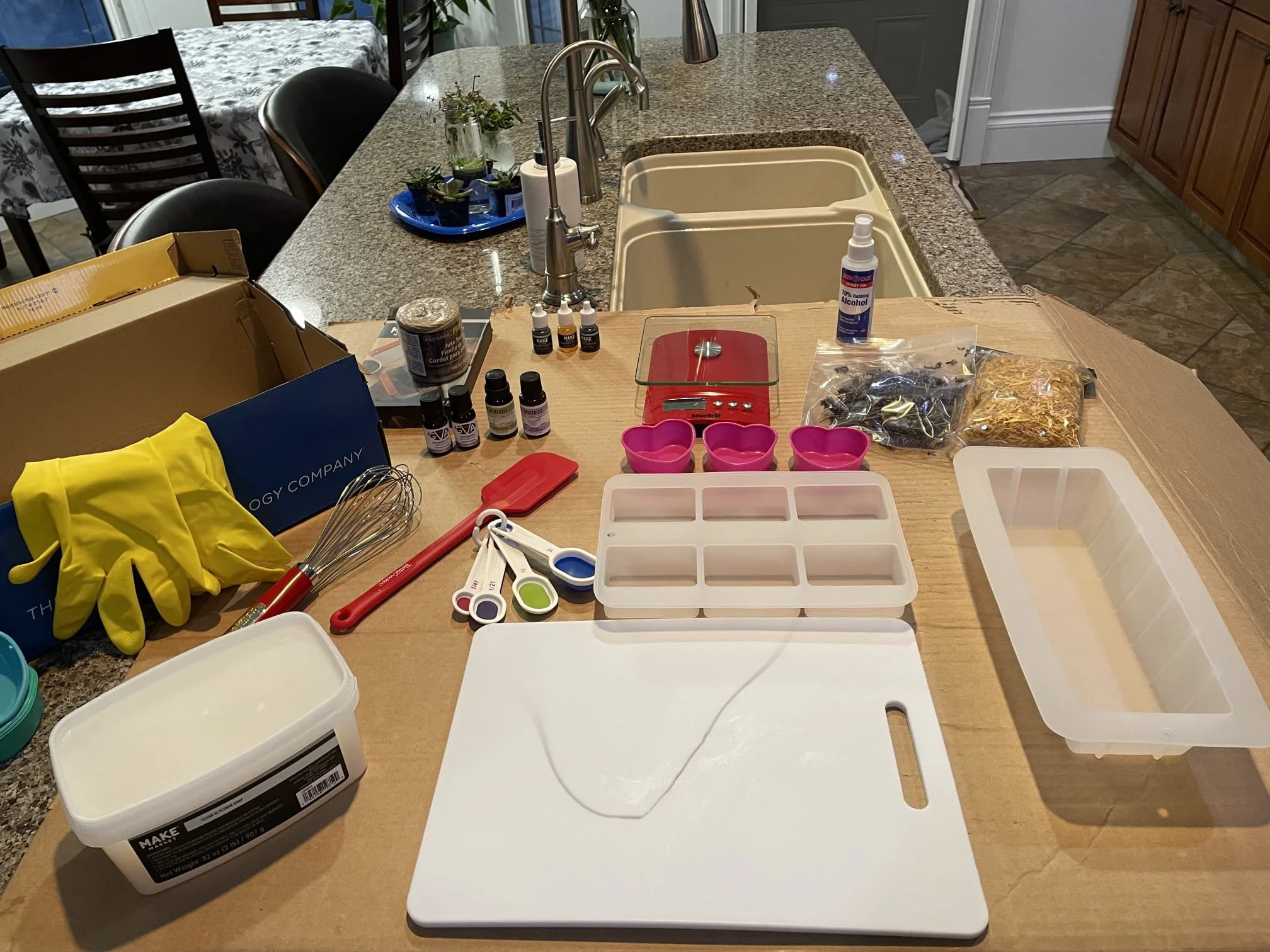 Kitchen counter with various baking supplies including gloves, measuring spoons, a cutting board, containers, a scale, flavor extracts, syrup, and packaged chocolate chips and nuts.