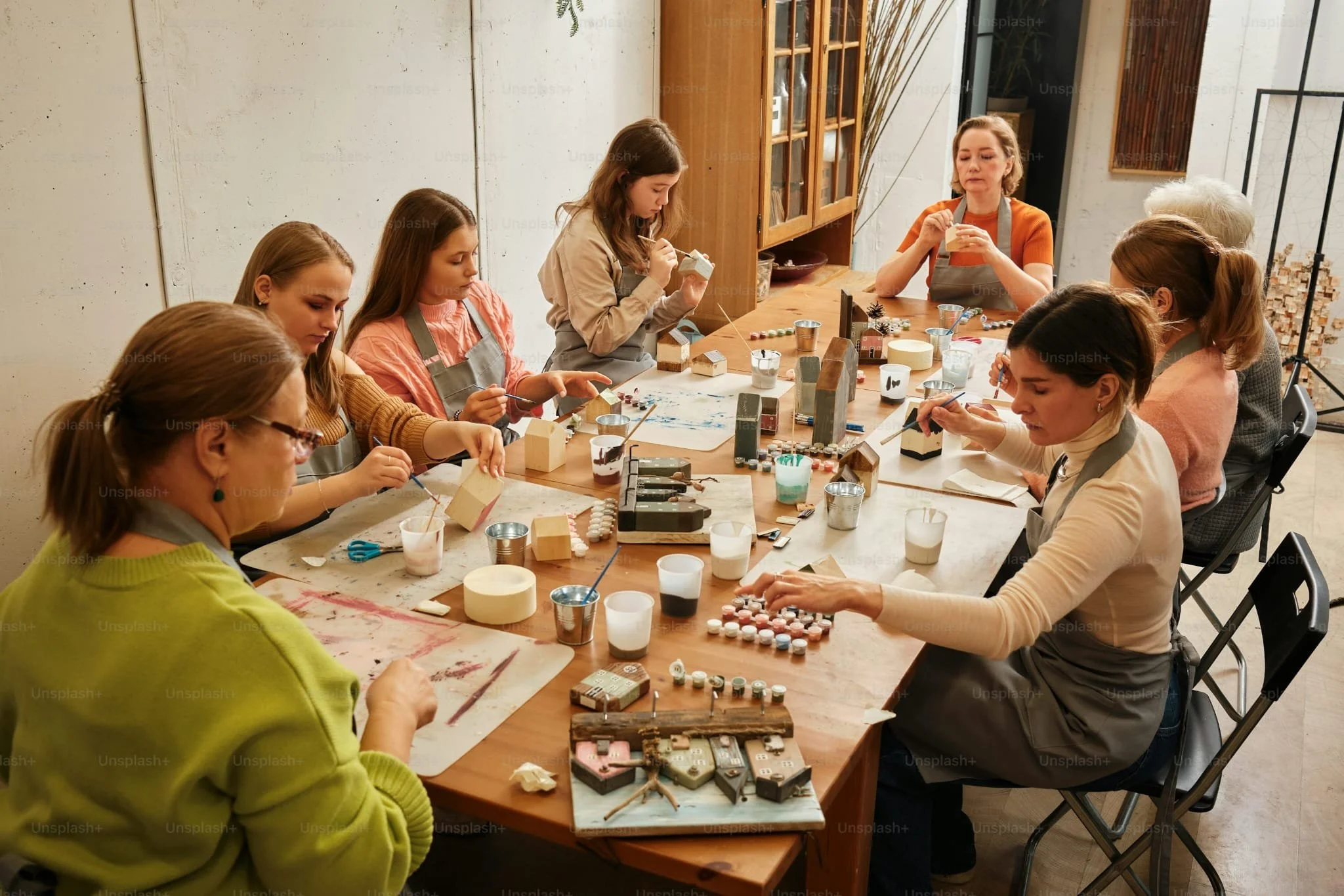 A group of women and girls practicing ceramic painting around a large wooden table in a cozy studio with art supplies, paint, brushes, and clay pieces.