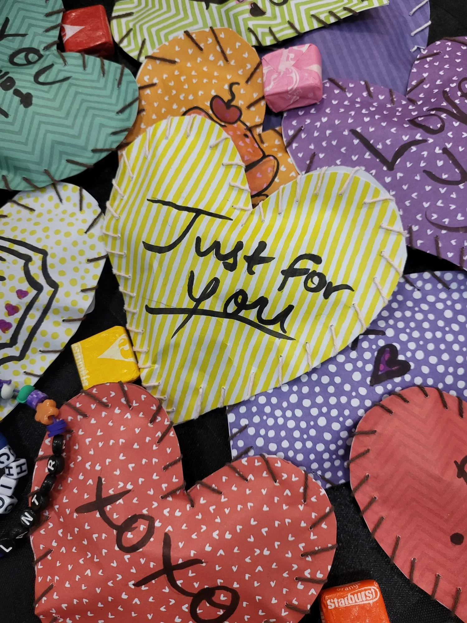 Colorful heart-shaped paper notes with messages, including one yellow heart with 'Just for You' written in black, surrounded by other hearts with words like 'Love' and decorated with small white and pink hearts, attached with black pins on a dark background.