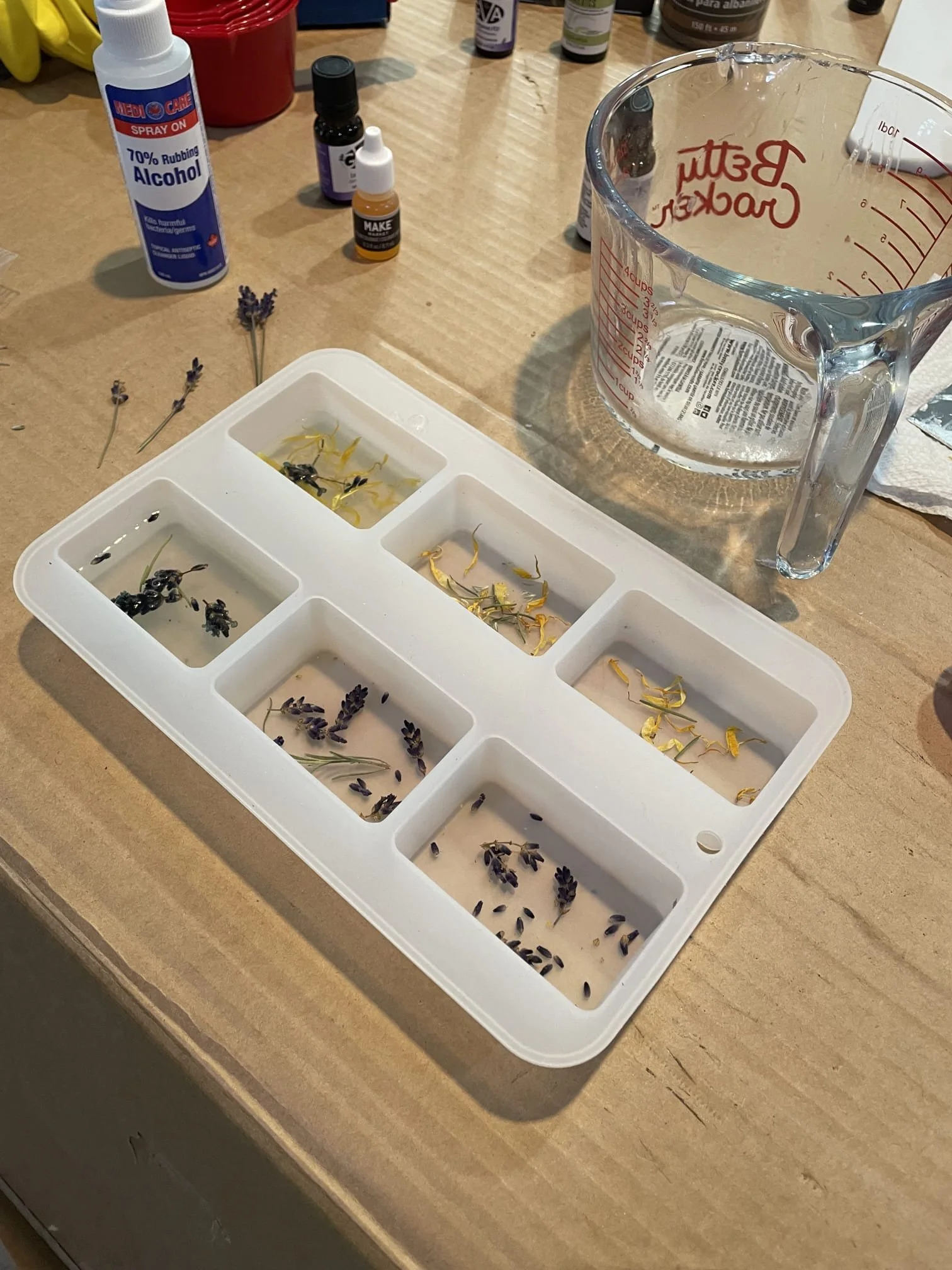 A white plastic tray with compartments containing small plant and flower specimens submerged in liquid, with a measuring cup, bottles of alcohol and essential oils, and sprigs of lavender on a wooden table.