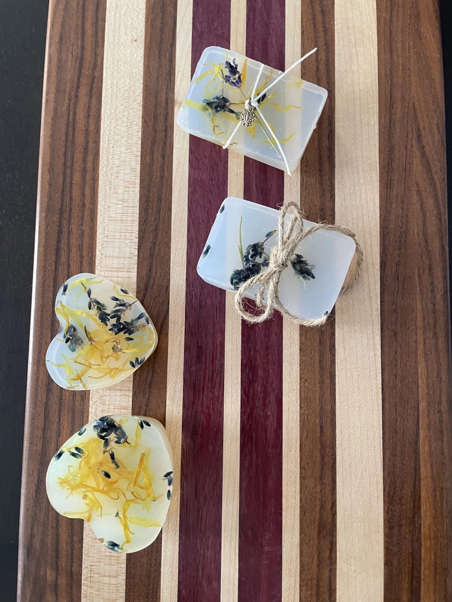 Four soap bars on a striped wooden surface, with three small soap bars containing yellow and black herbs, and one larger white soap bar with a silver charm and twine, and another white soap with herbs.
