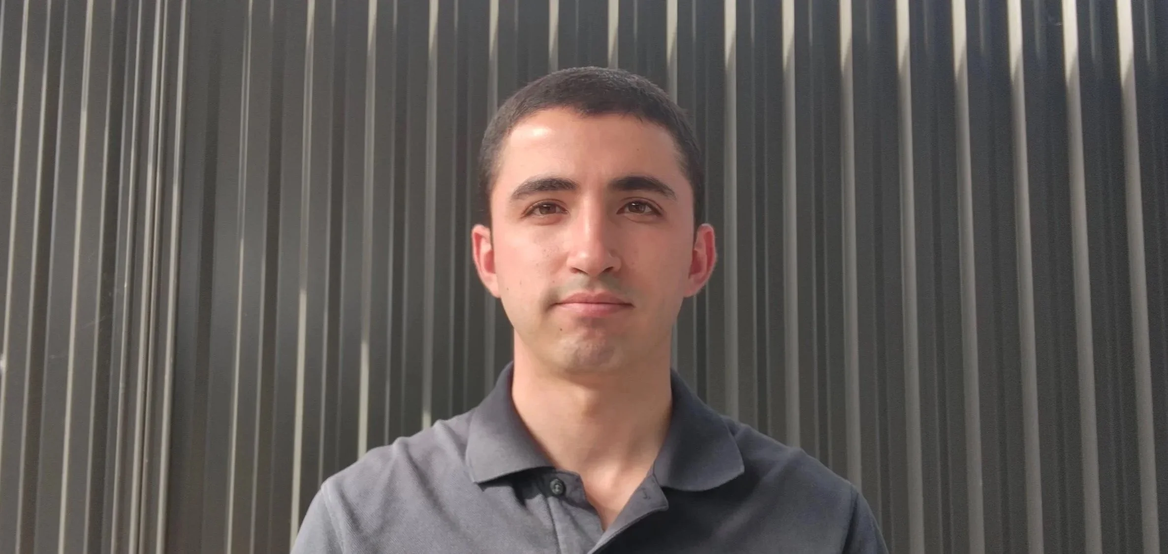 Close-up of a young man with short dark hair, wearing a blue polo shirt, standing in front of a dark, vertical corrugated metal wall.