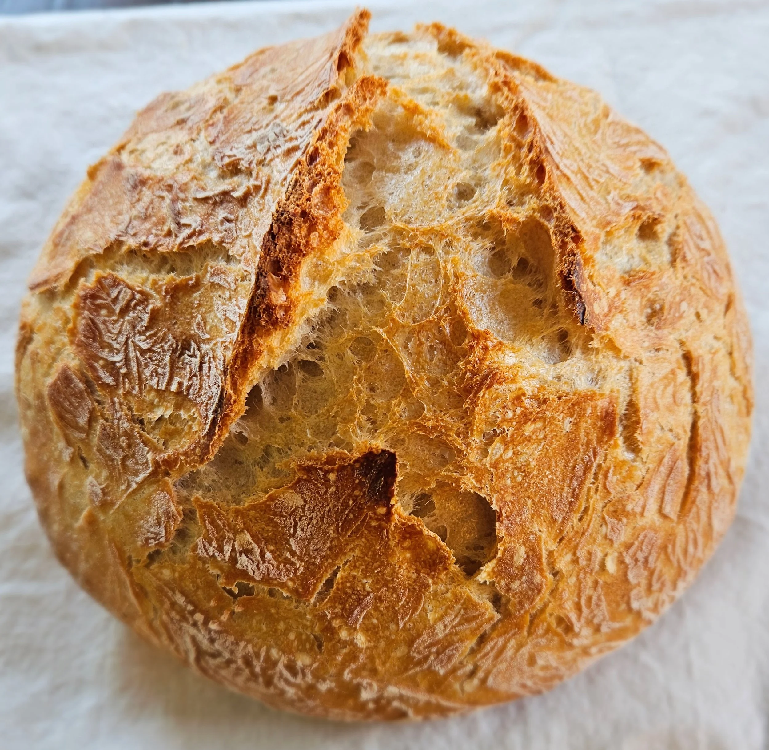 Close-up of a freshly baked Artisan White Boule with a deeply scored, golden-brown crust showing an open, irregular crumb texture, resting on a white linen surface.