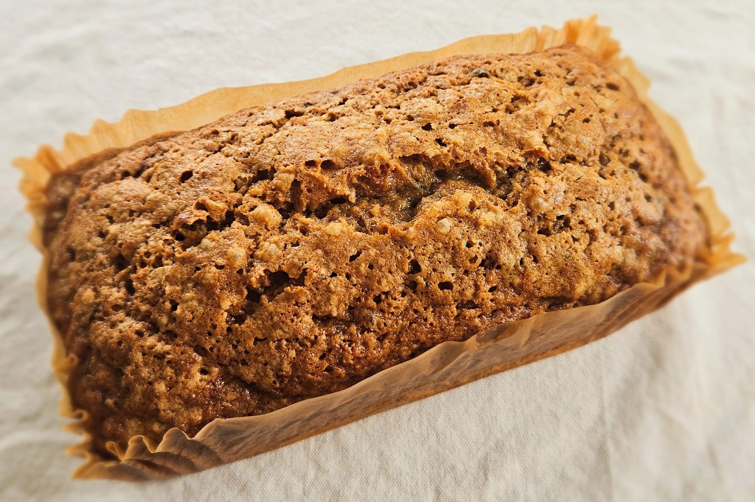 Whole Zucchini Walnut Loaf in its parchment liner resting on a white linen surface, showing the rustic, cracked top crust with a warm amber-brown color.