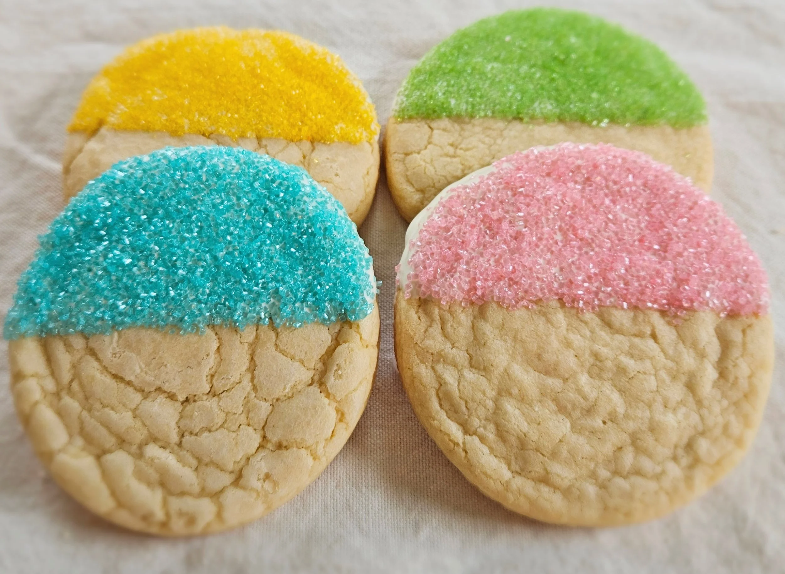 Four Half-Dipped Sugar Cookies arranged in a cluster on white linen showing all four pastel colors — yellow, green, blue, and pink — each half-dipped in white chocolate with sparkling sanding sugar.