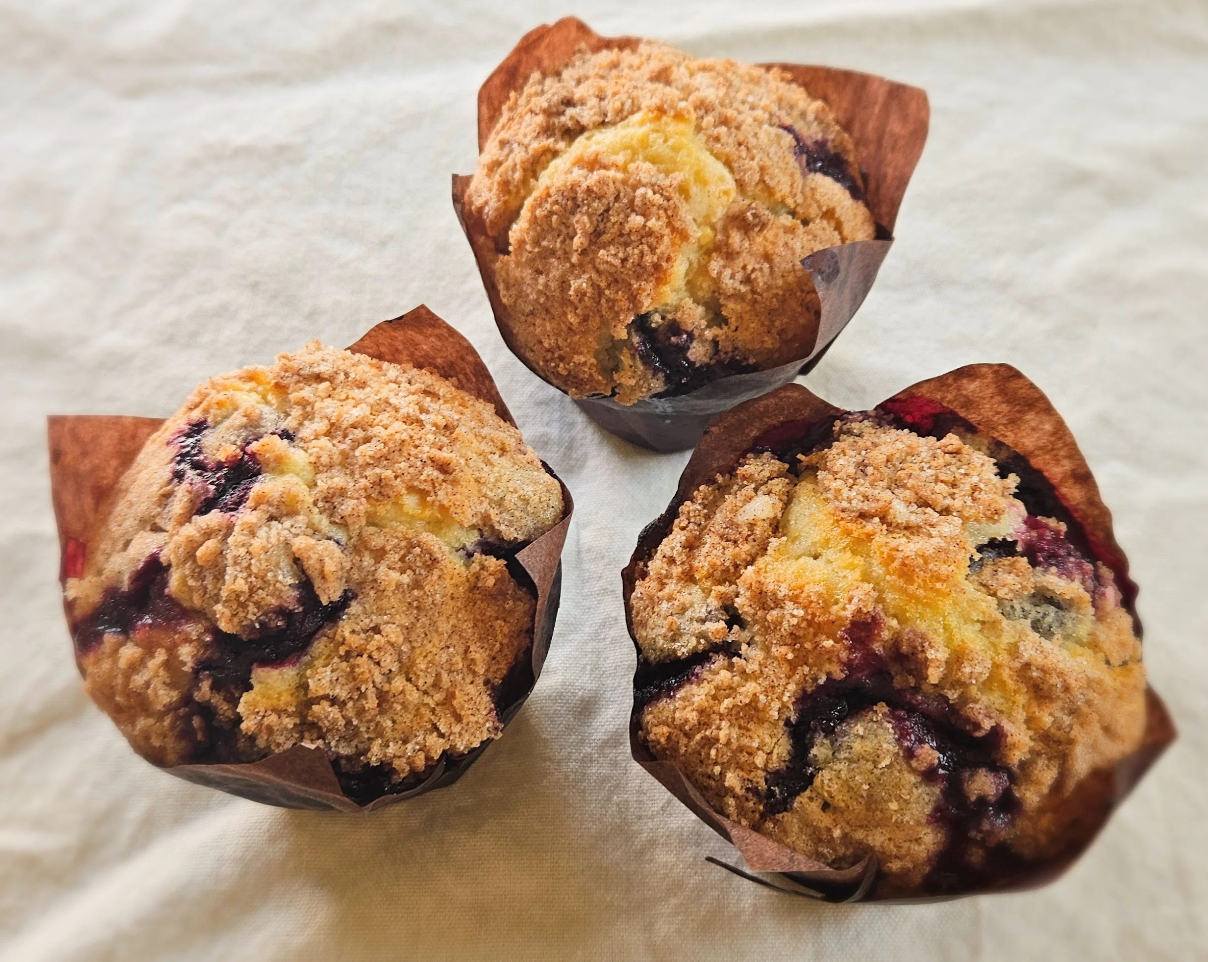 Three oversized Blueberry Crumble Muffins in brown tulip liners arranged in a triangle on a white linen surface, showing golden crumble topping and blueberries bursting through the muffin sides.