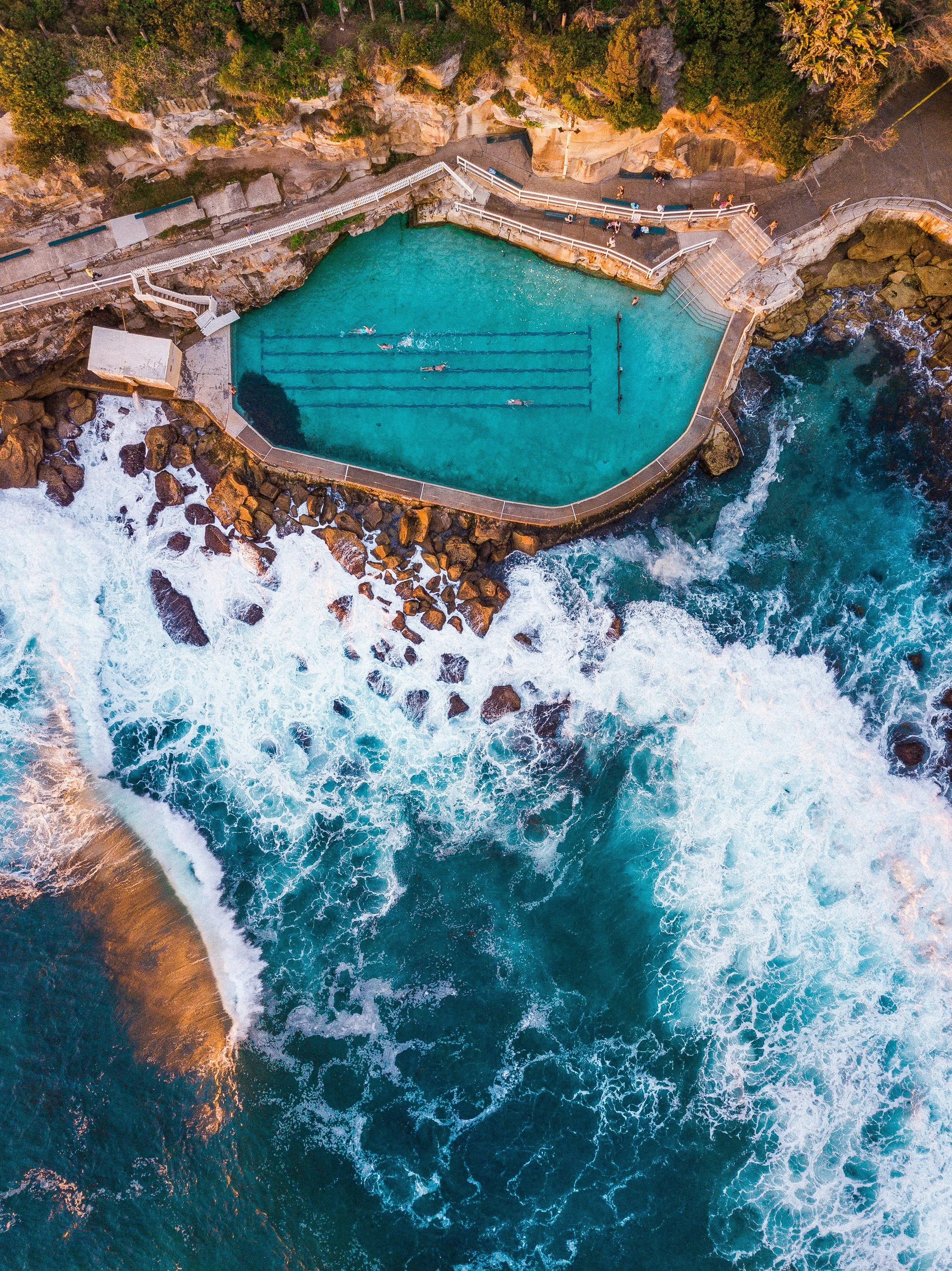 An aerial view of a scenic coastal infinity pool with swimmers, surrounded by rocky cliffs and lush greenery, overlooking the ocean.