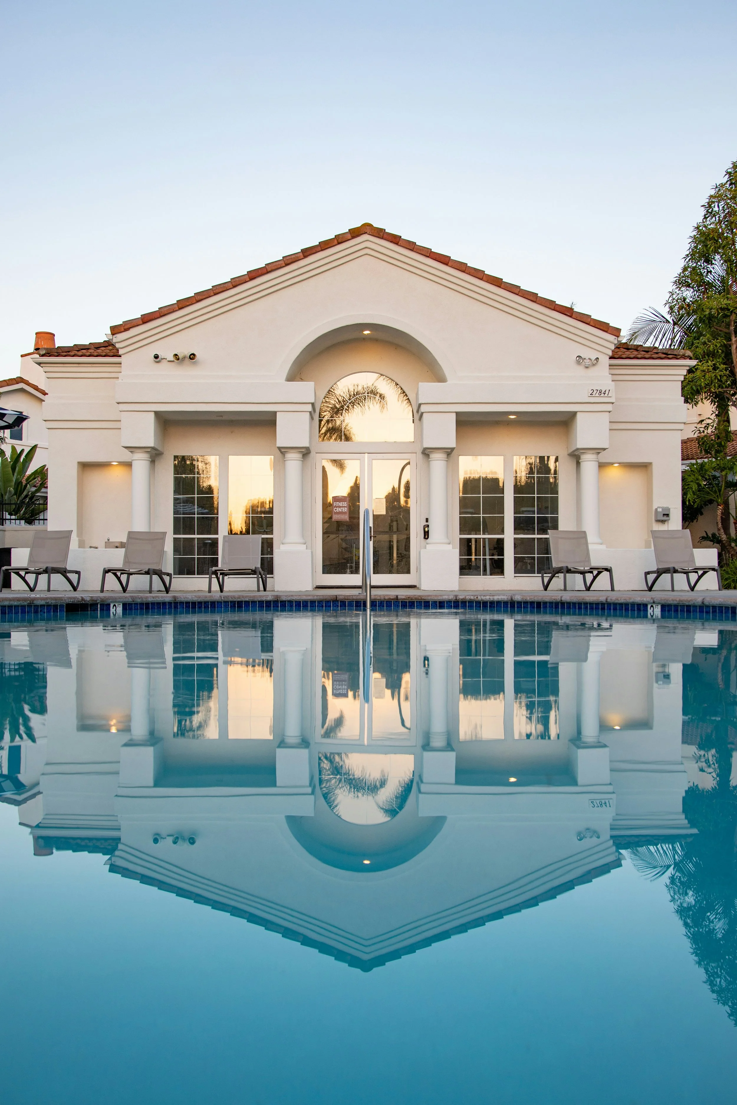 A white building with large windows and a red-tiled roof, reflected in a swimming pool in the foreground, with lounge chairs along the poolside.