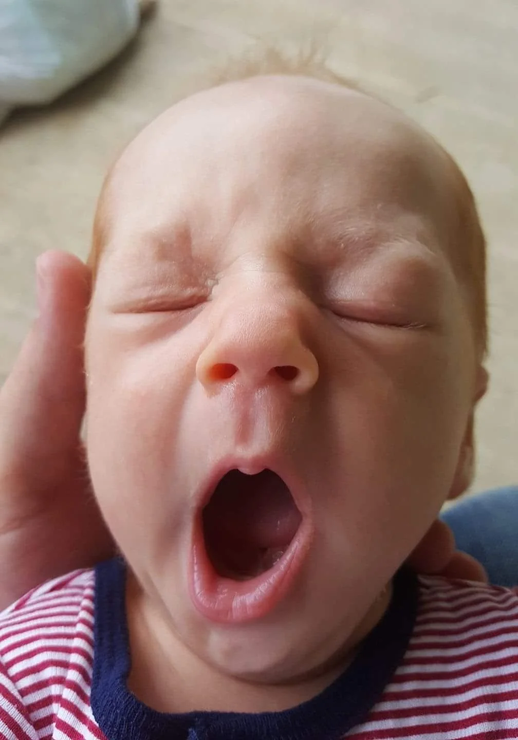 Close-up of a yawning baby with closed eyes and open mouth, wearing a red and white striped shirt.