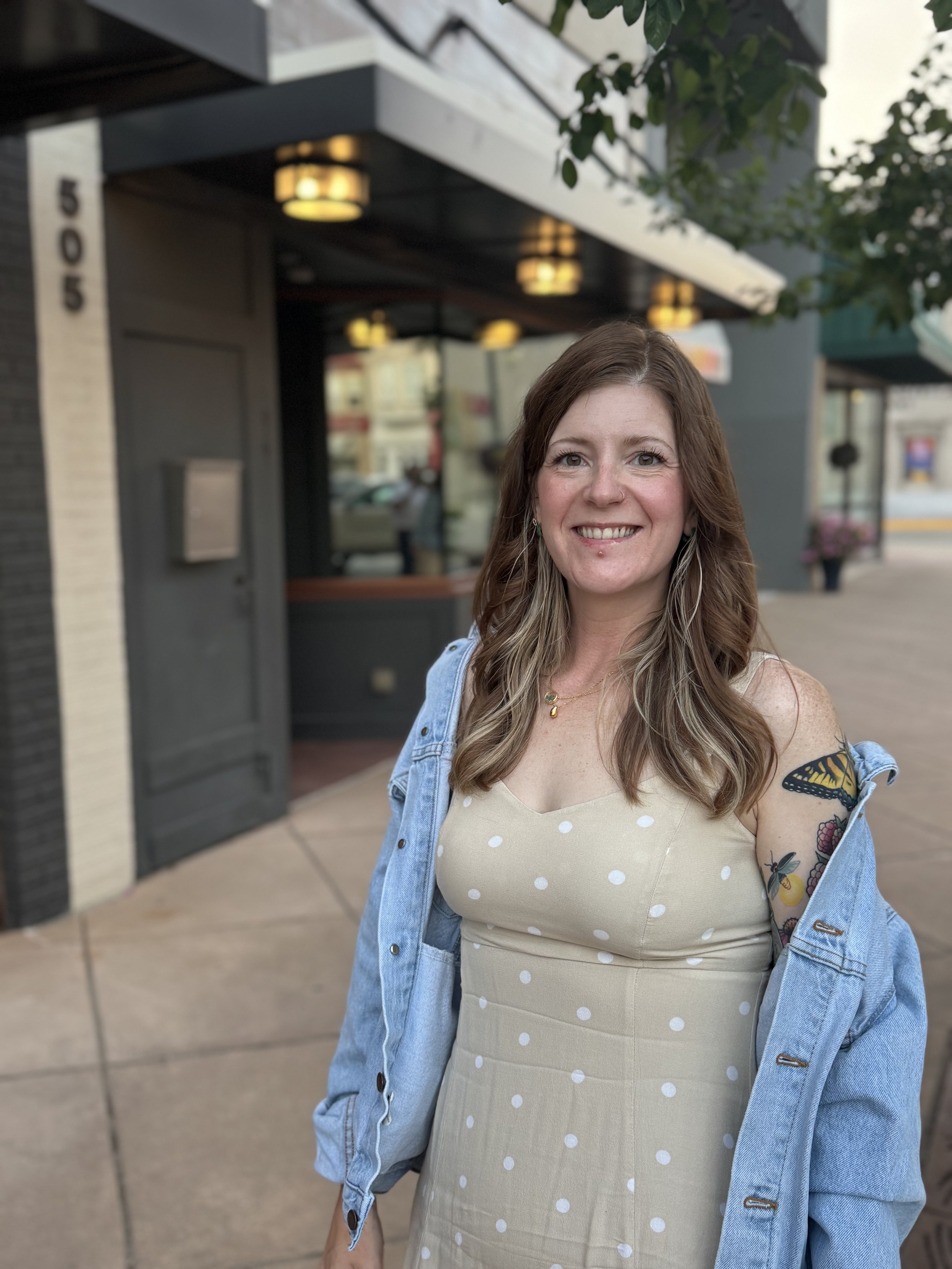 A woman with brown hair smiling outdoors, wearing a beige polka dot dress and a light blue denim jacket, standing on a sidewalk in front of a building with the number 505.