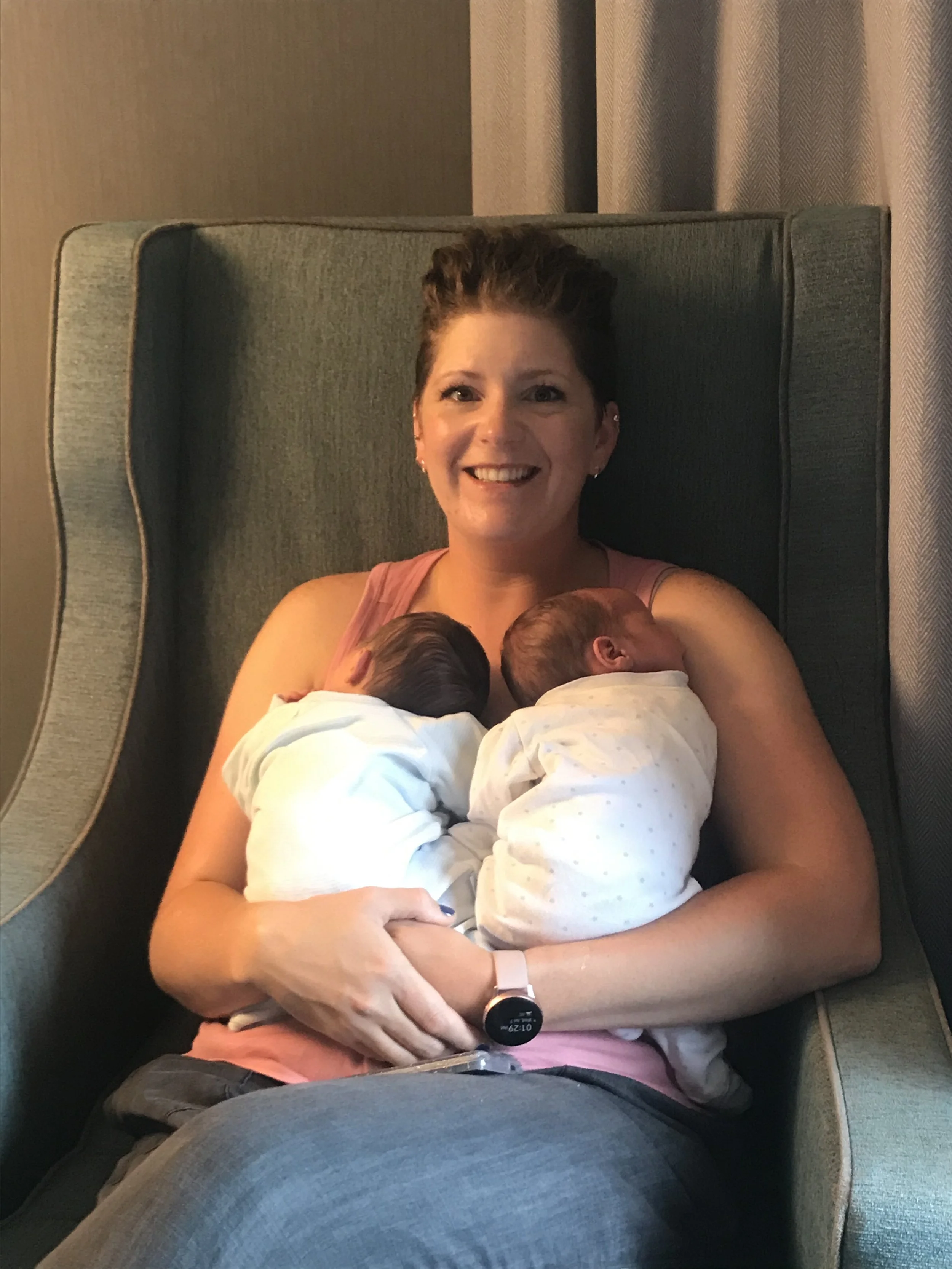 A woman smiling while breastfeeding two newborn babies, sitting in a teal armchair with curtains behind her.