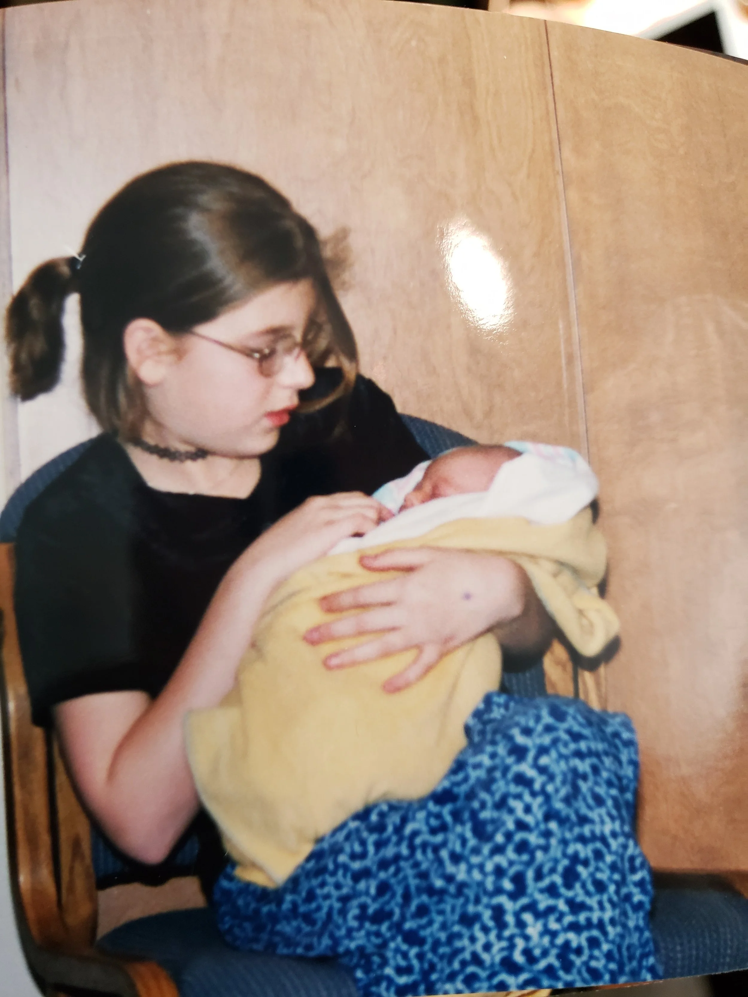 A young woman with glasses and dark hair pulled back in a ponytail holding and looking at a newborn baby wrapped in a yellow blanket.