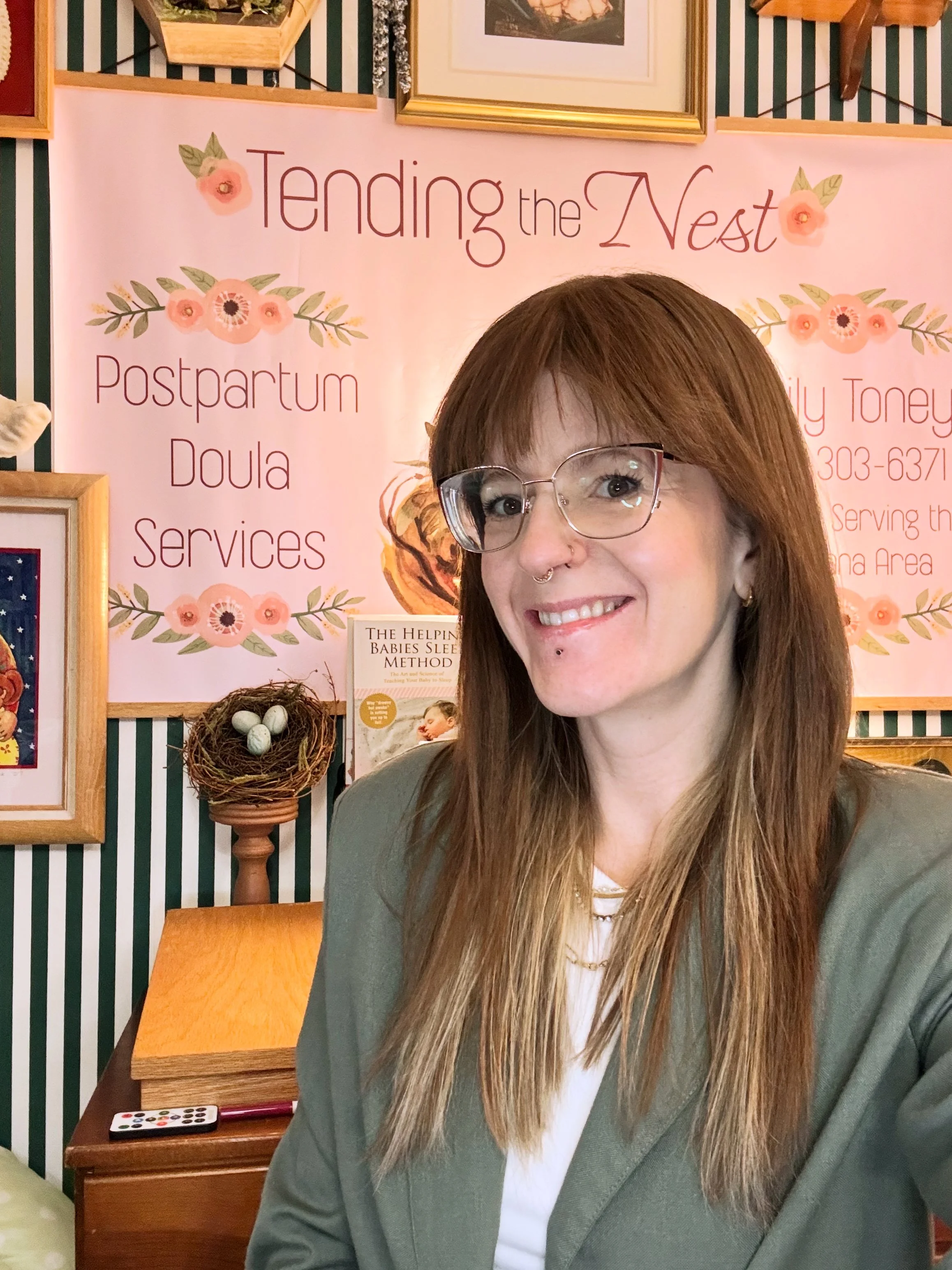 A woman with long brown hair, glasses, and a septum piercing, smiling and taking a selfie in front of a pink floral sign that reads 'Tending the Nest Postpartum Doula Services'.