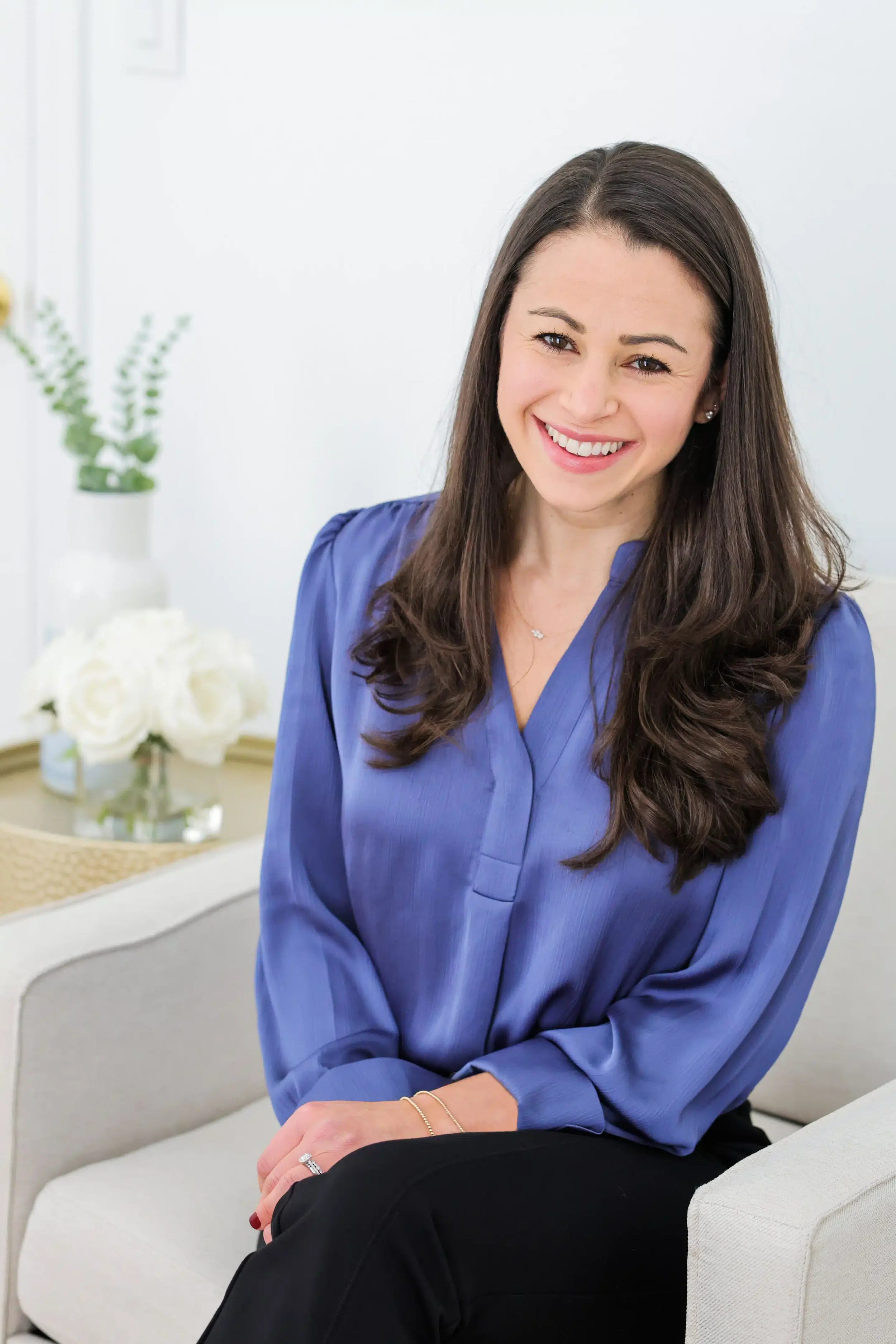 A woman with long brown hair wearing a blue blouse and black pants sitting on a beige chair in a bright space with white walls, a white vase with flowers, and a small table with a glass of water in the background.
