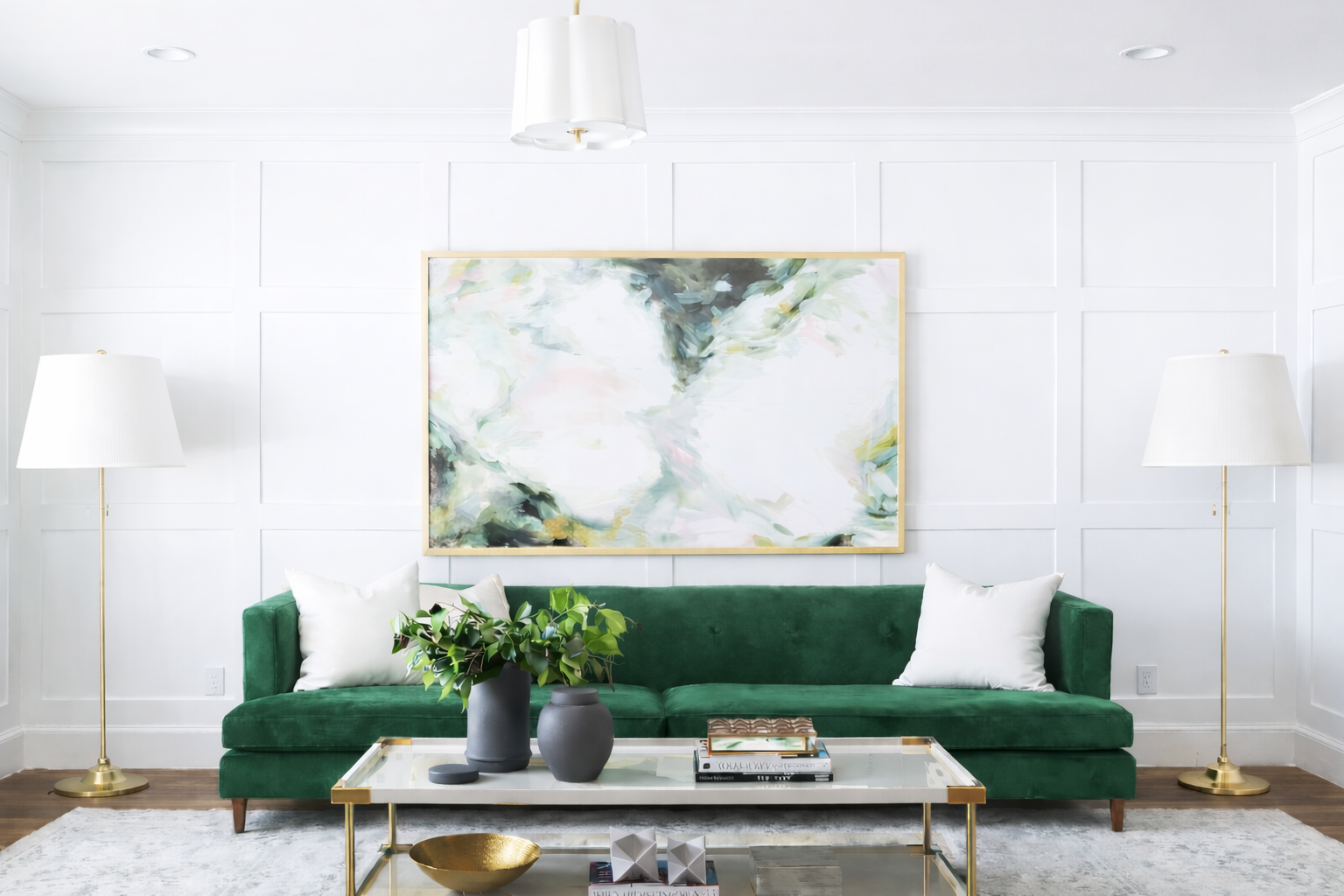 Living room with white walls, a green sofa, two white pillows, a glass coffee table with pots of greenery, and two white floor lamps on gold stands. Above the sofa is an abstract painting.