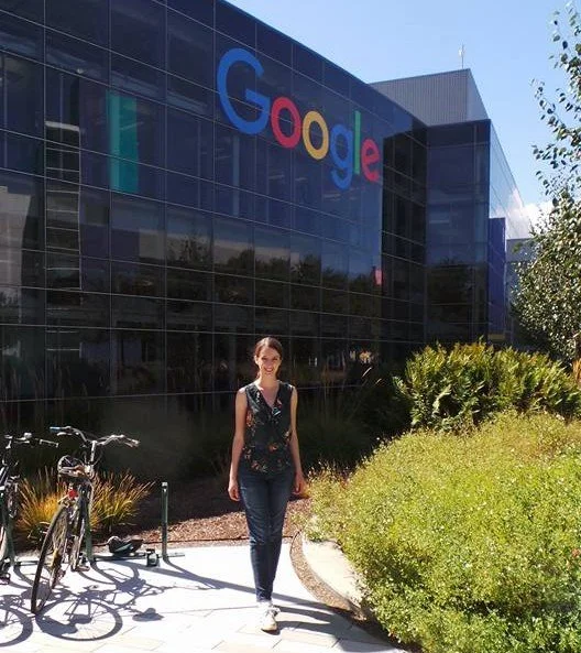 Eilonwy Kennedy walking outside the Google headquarters in San Francisco, which has a large glass building with the Google logo on it, and bicycles parked nearby.
