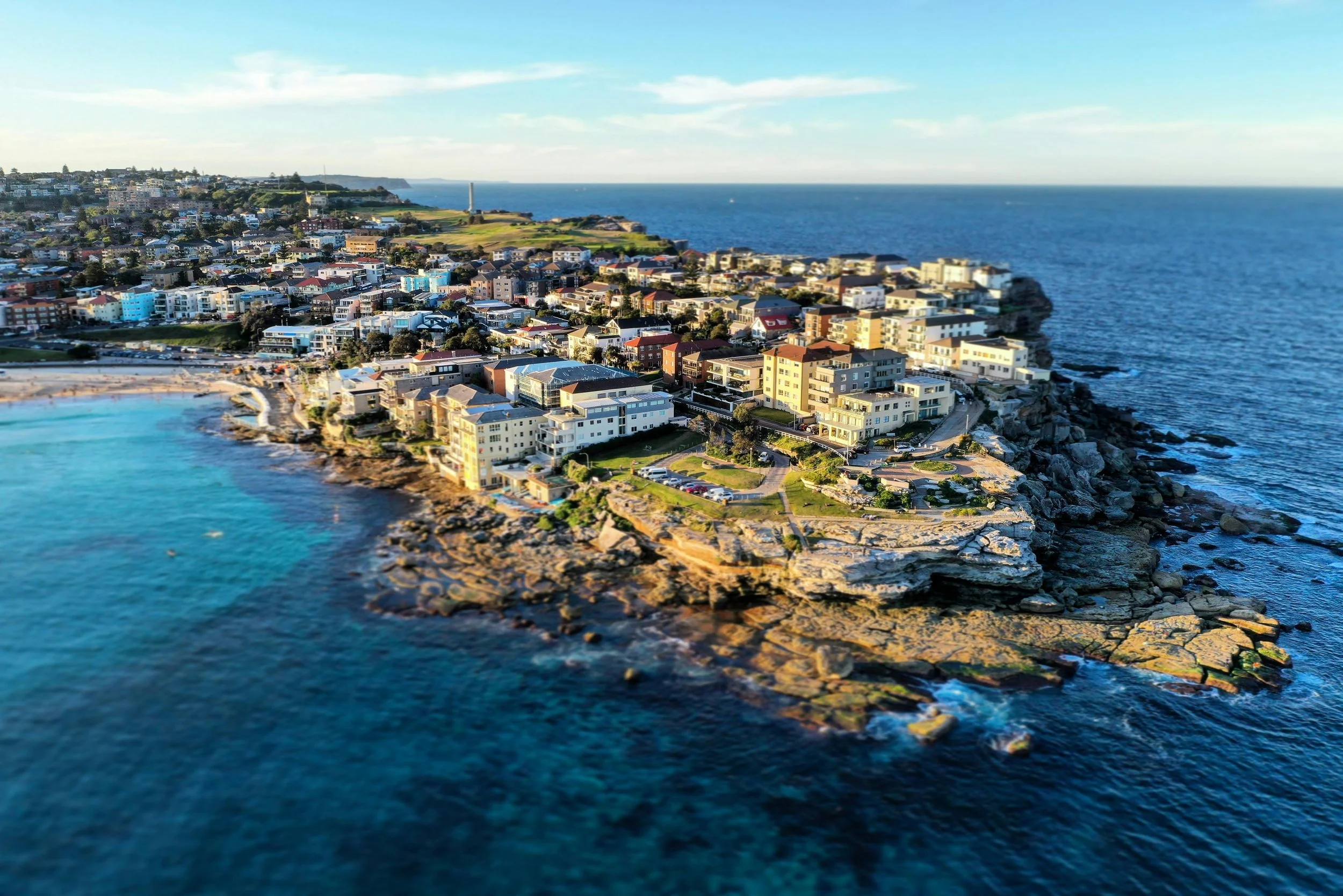 Aerial view of Bondi beach with multicolored buildings, rocky shoreline, and deep blue ocean under a partly cloudy sky.