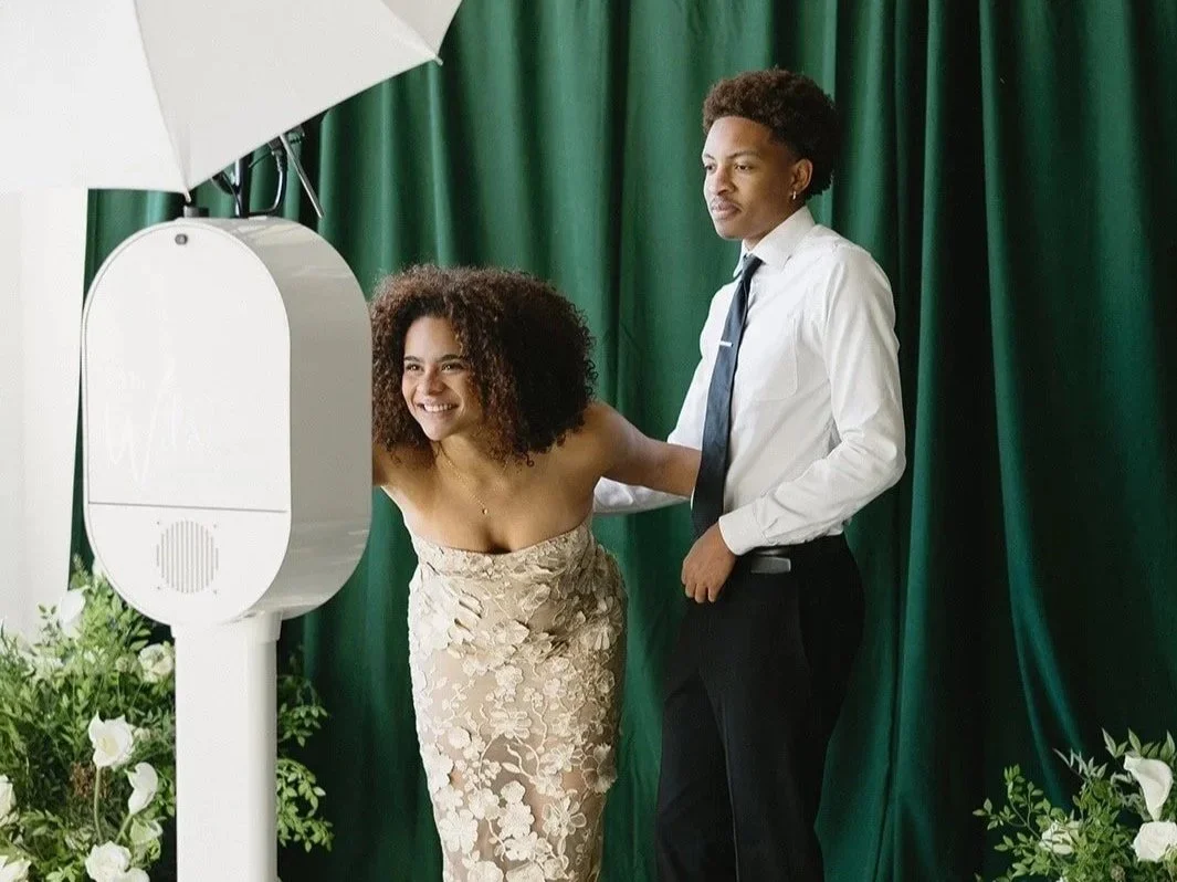 Two people posing for a photo in a photo booth, with a green curtain background and a white umbrella reflector, one woman smiling and leaning in, the man standing next to her.