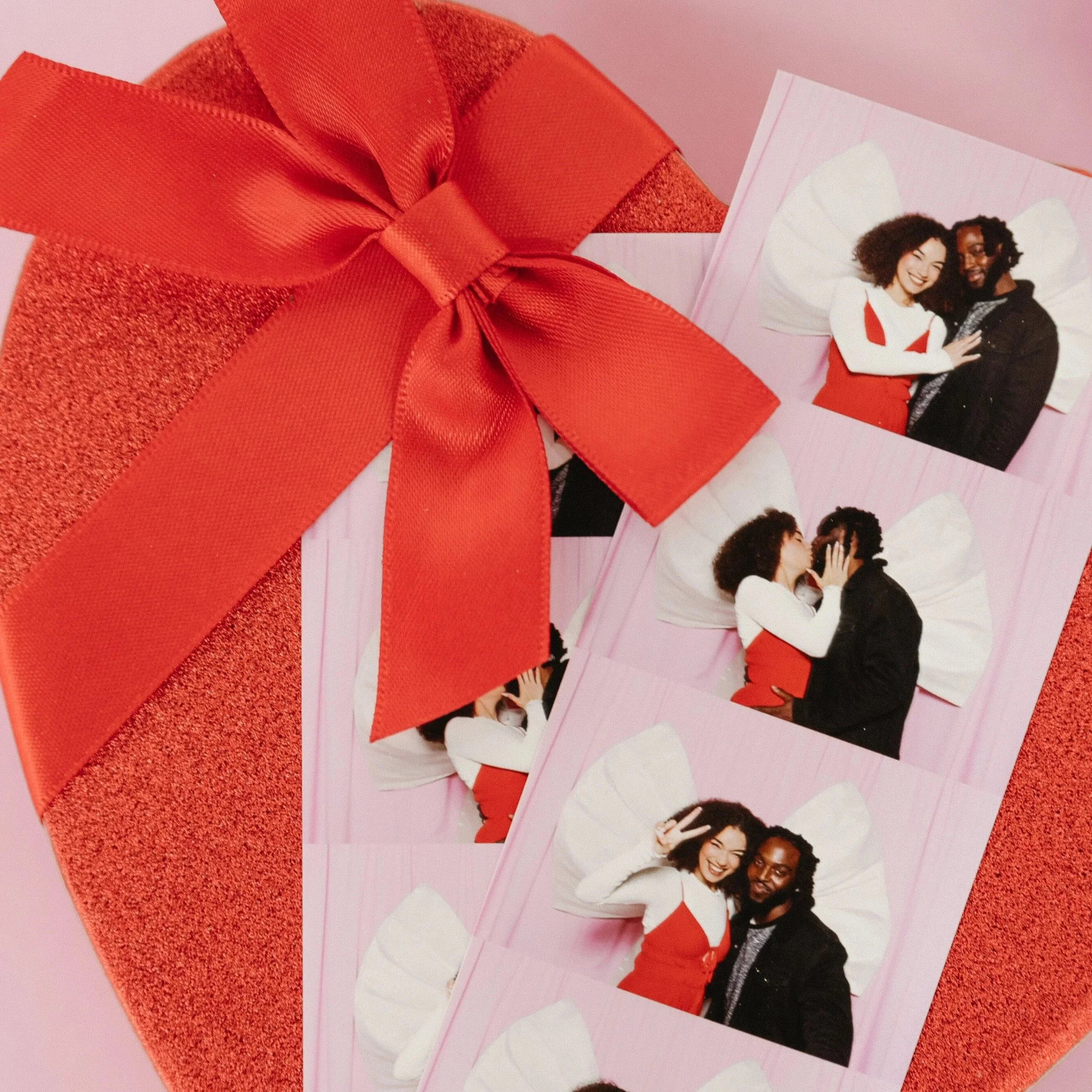 A red gift box with a wide satin ribbon bow on top, placed on a pink surface. Next to it are photo booth strips showing a couple with butterfly wings, taking playful pictures together.