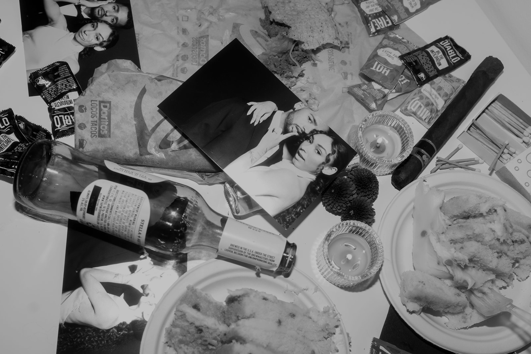 A table with partially eaten plates of cake, scattered snack packages including Doritos and Snickers, a partially crushed glass bottle, and photos of a man and woman. There are lit candles, small decorative items, and miscellaneous objects on the table.