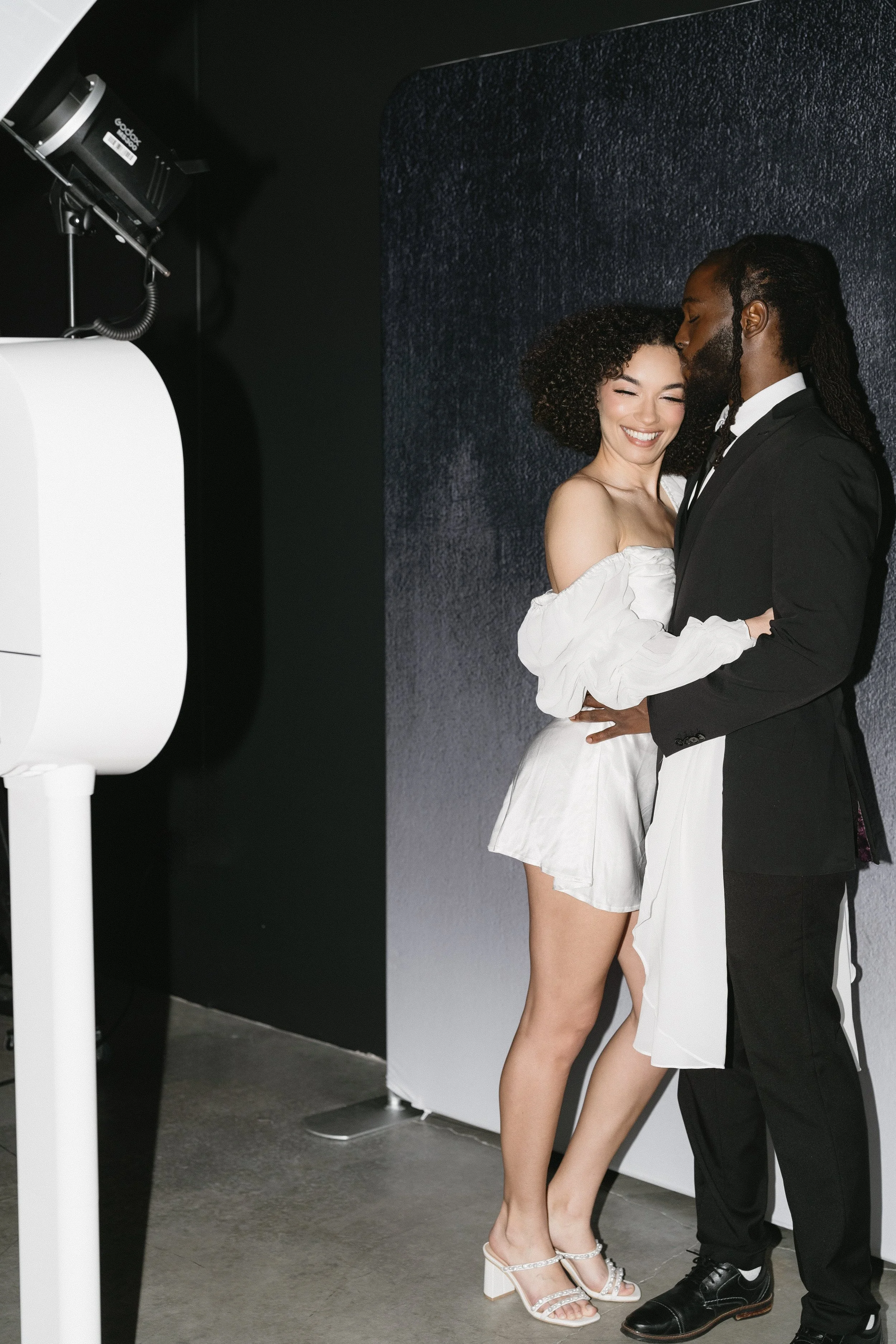A couple dressed in formal wedding attire posing and embracing together, smiling, in front of a dark backdrop with professional lighting nearby.