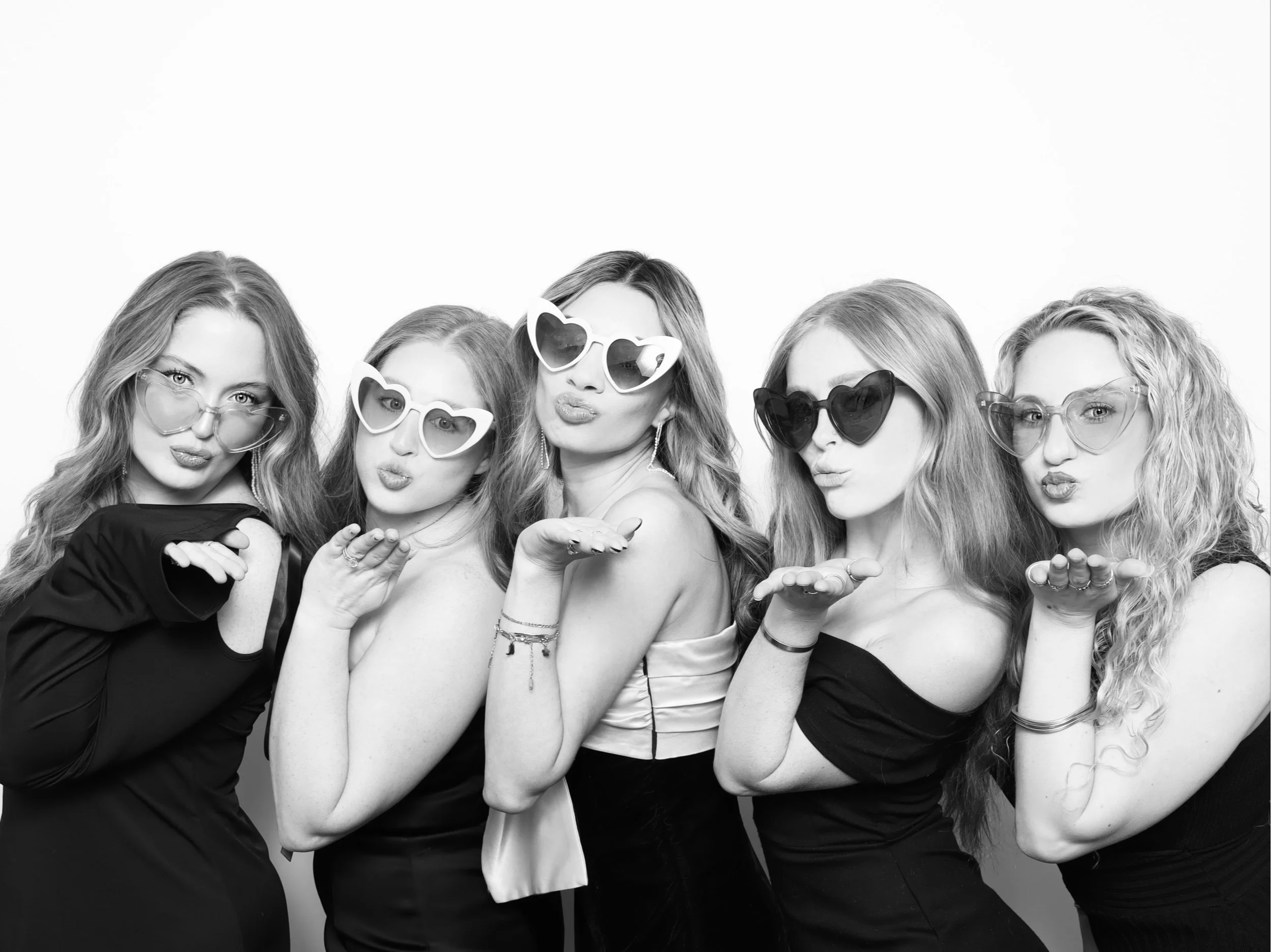 Five women wearing stylish sunglasses blowing kisses towards the camera in a black-and-white photo.