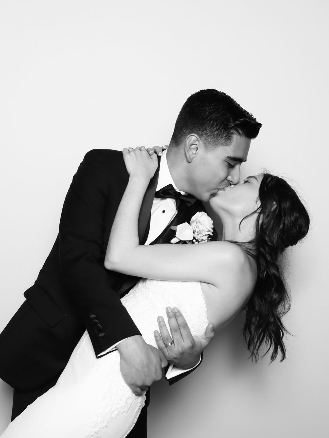 A black-and-white photo of a couple in wedding attire sharing a kiss, with the man wearing a tuxedo and the woman in a strapless wedding dress.