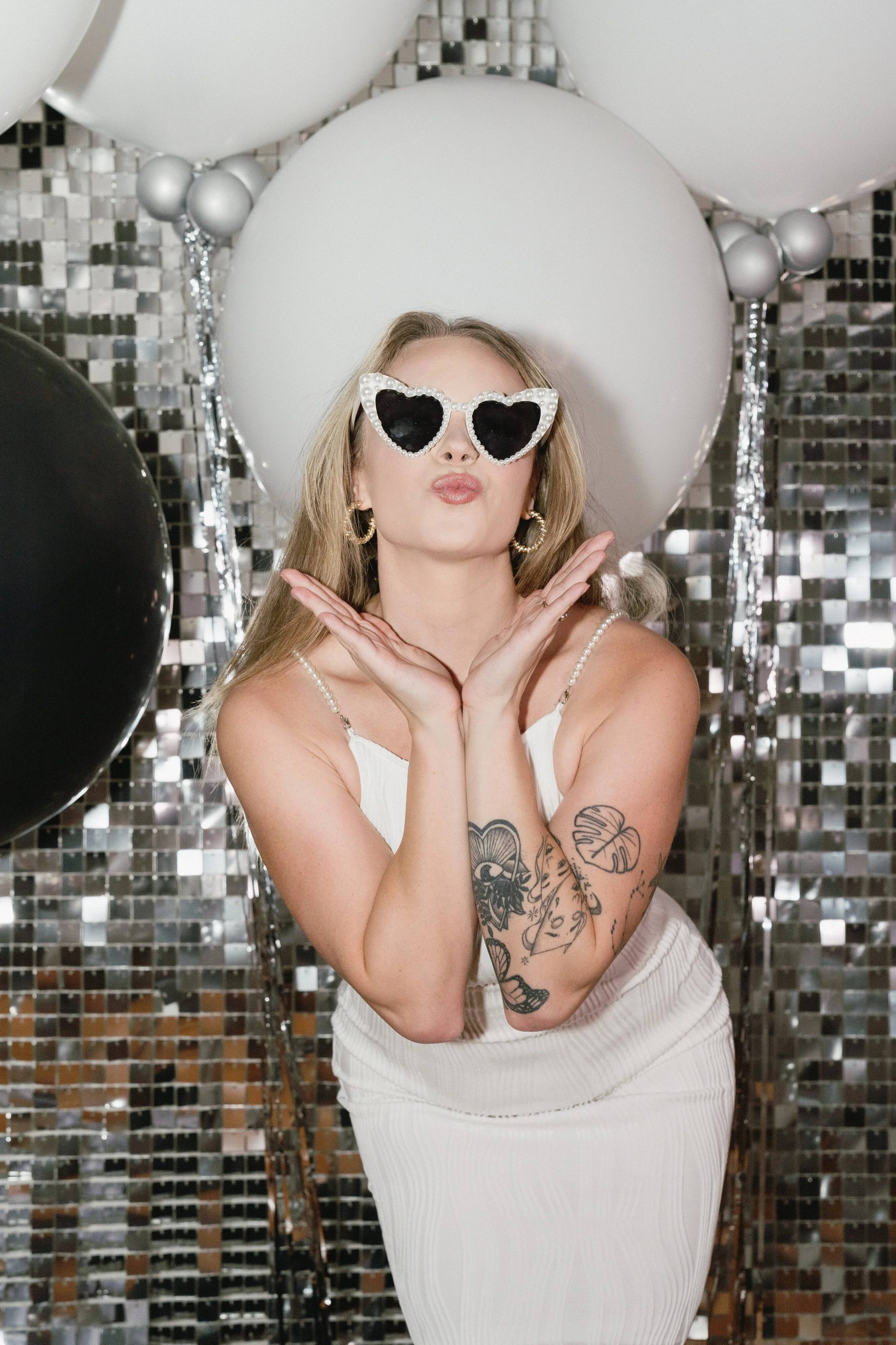 A woman with blonde hair wearing heart-shaped sunglasses, earrings, and a white dress, is posing with her hands near her face at a party with balloons and a mirrored backdrop.