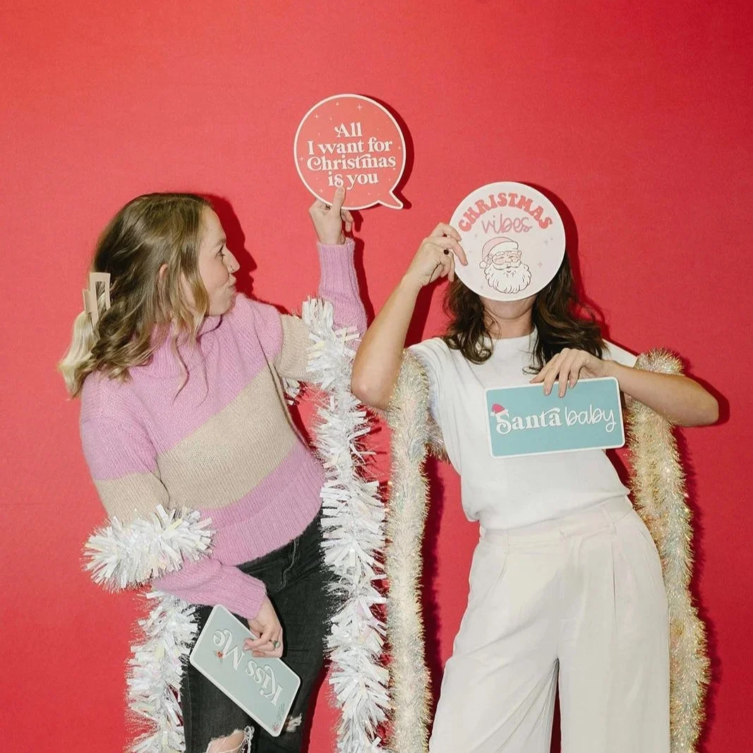 Two women with Christmas-themed signs and accessories stand against a red background. One woman holds a sign that says 'All I want for Christmas is you.' The other holds a sign that says 'Santa baby' and a round sign with a Santa face that says 'Christmas vibes.' They are dressed festively with one wearing a pink and beige sweater and the other a white outfit with gold tinsel.