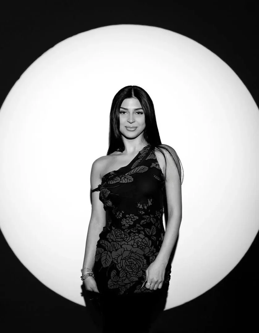 A woman with long dark hair wearing a black dress with floral pattern poses in front of a large circular light source creating a halo effect.