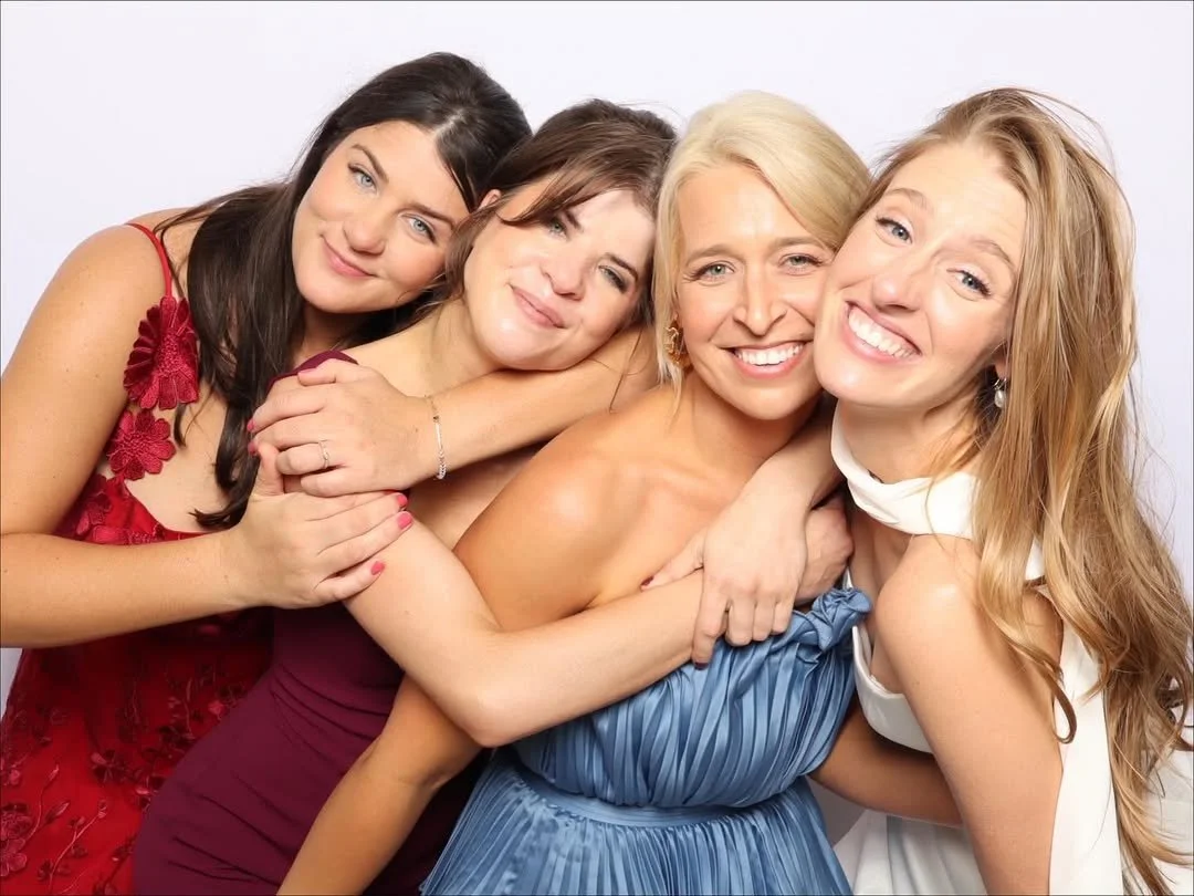 Four women dressed in elegant gowns are embracing and smiling at the camera against a plain light background.