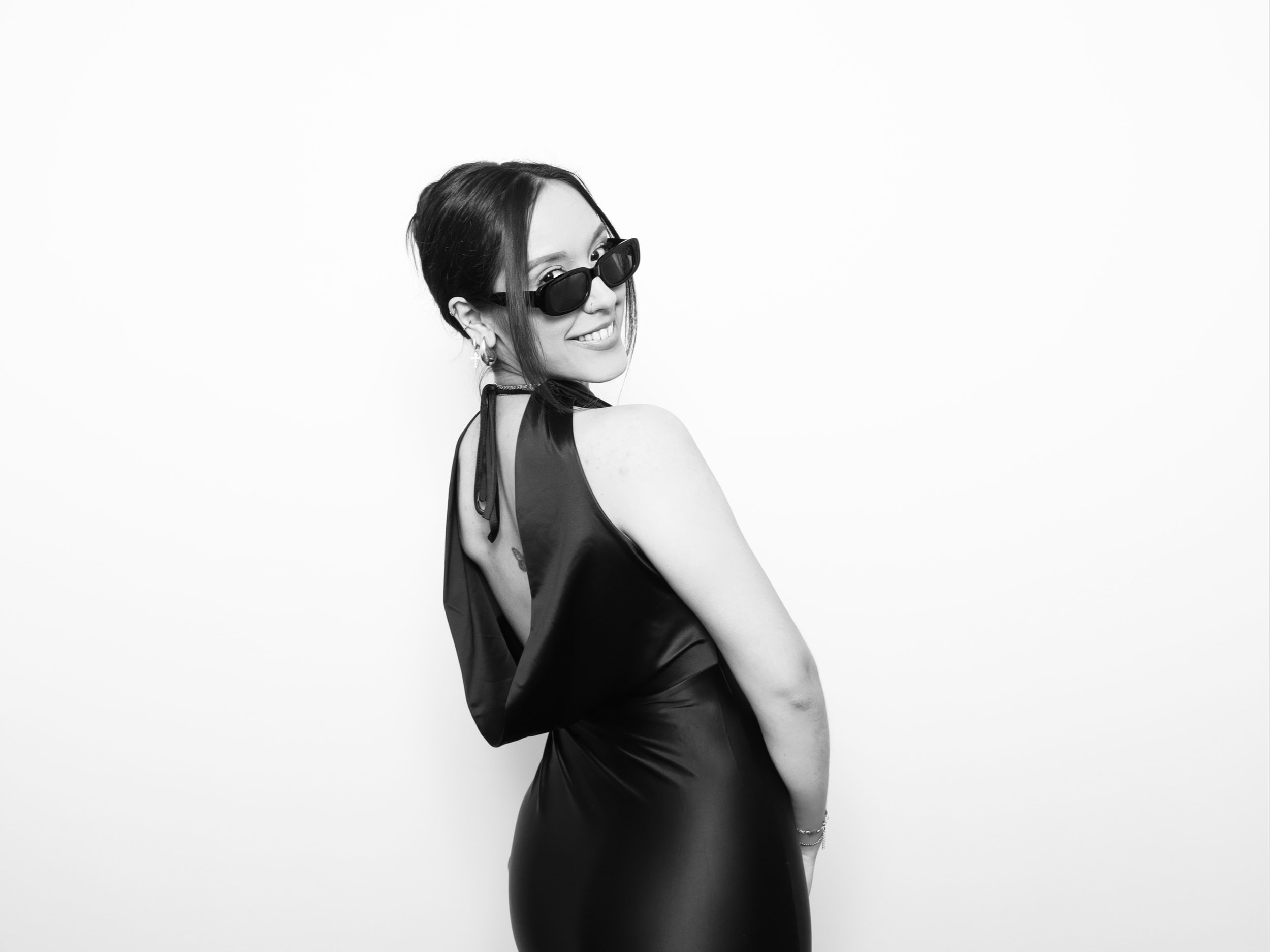 Black and white photo of a woman with dark hair in a partly updo, wearing sunglasses, a satin halter top, and jewelry, smiling and looking over her shoulder against a plain white background.