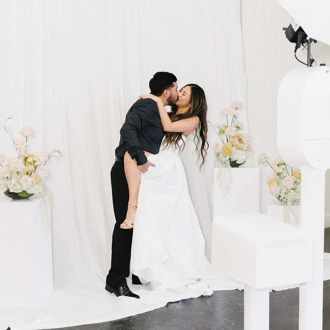 A couple kissing during a wedding photo shoot, with the groom lifting the bride in a white wedding dress and the groom dressed in black, surrounded by flower arrangements and a white backdrop.