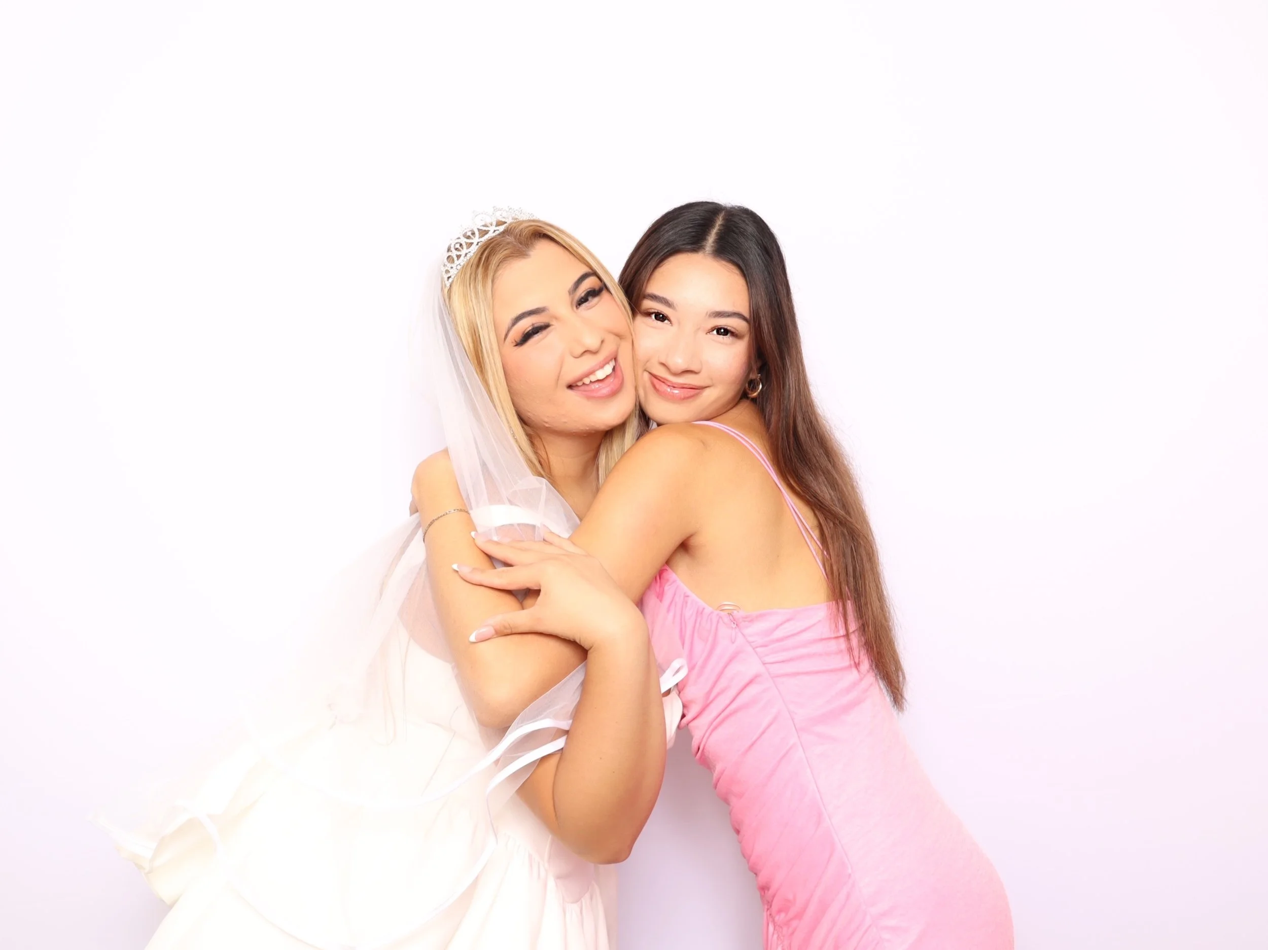 Two young women hugging and smiling, one dressed as a bride with a veil and tiara, and the other in a pink dress, posing against a white background.