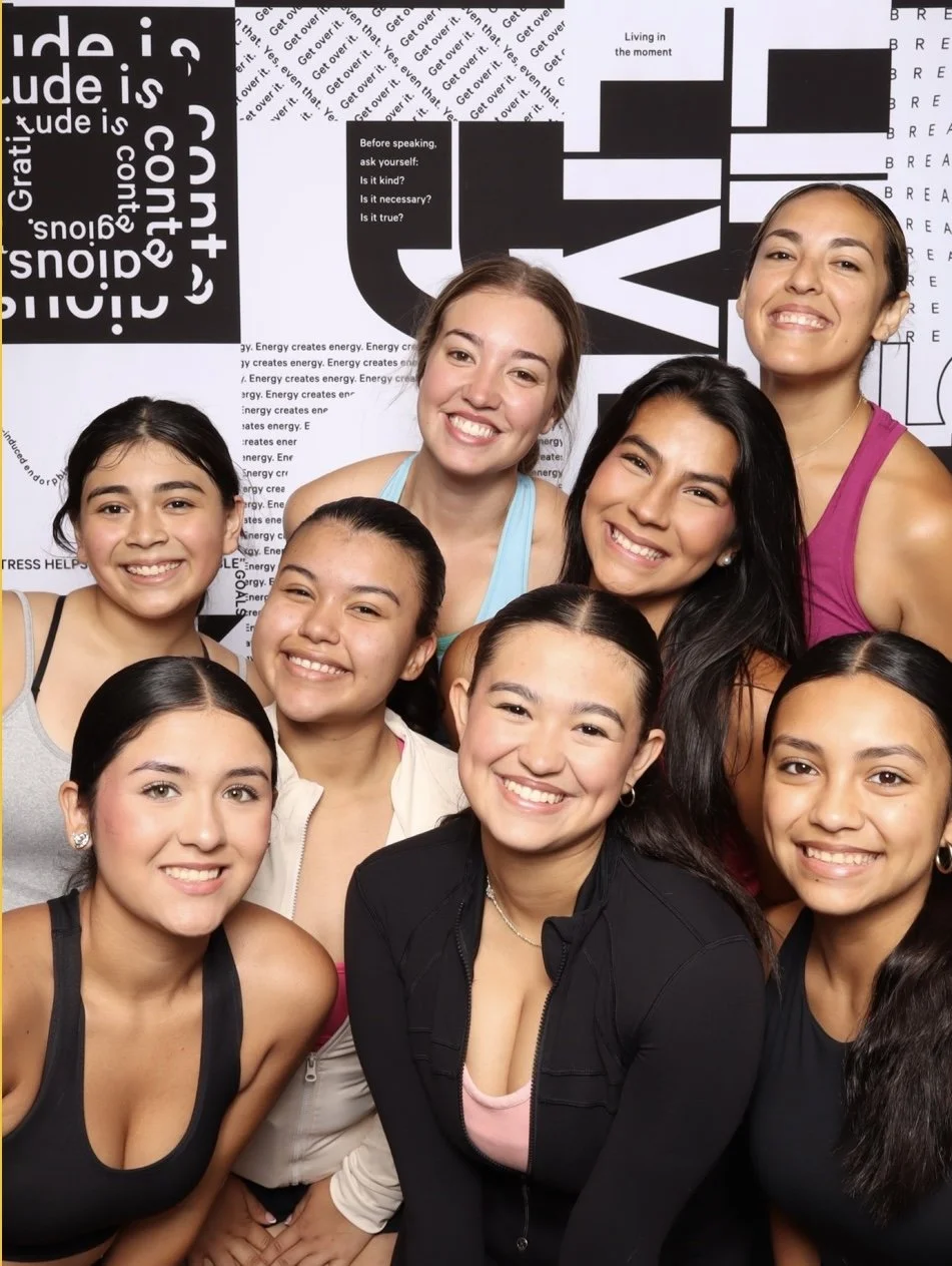 A group of nine women smiling and posing together in front of a black and white abstract background with text about attitude, living in the moment, and energy creation.