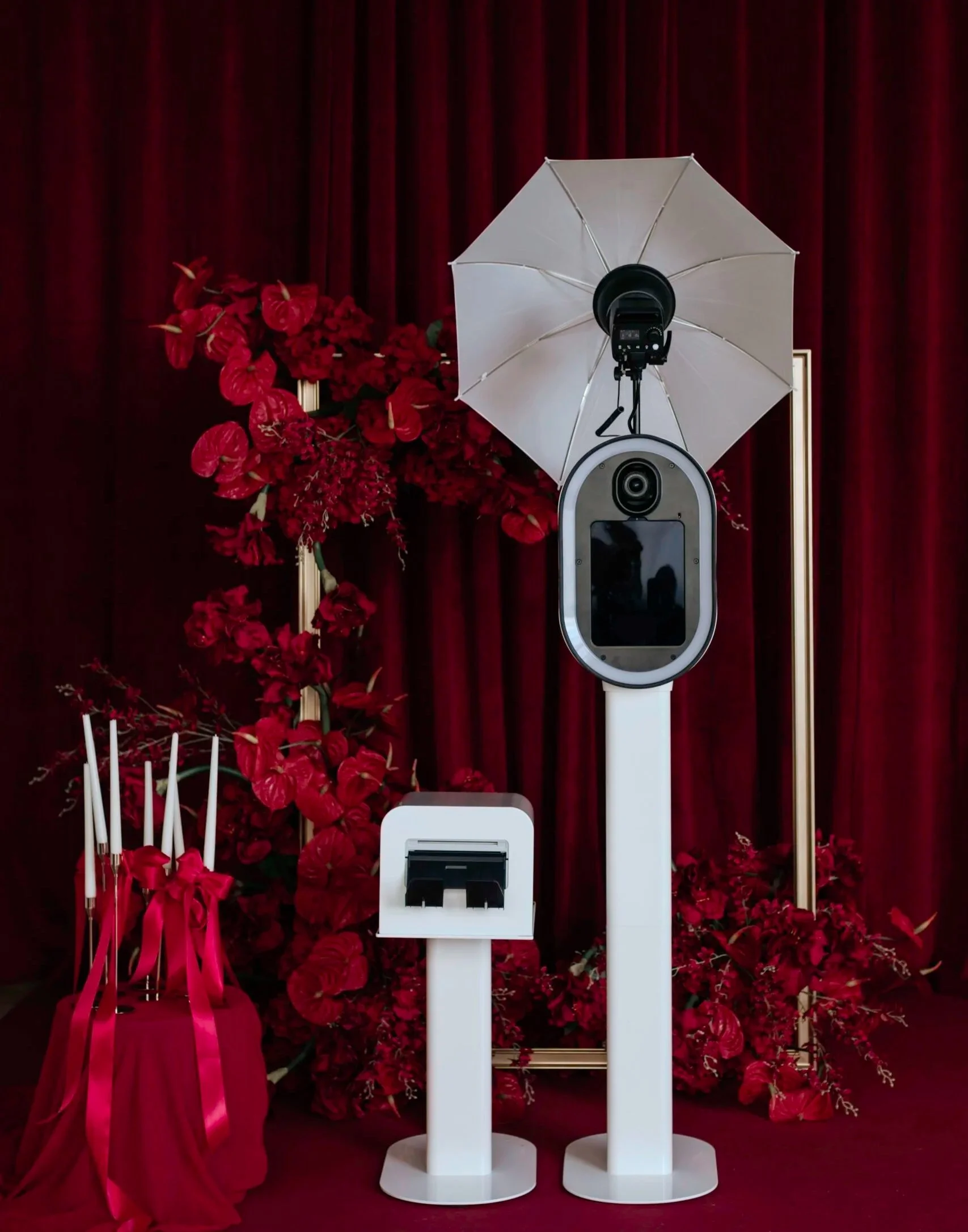 A photo of a photobooth setup with a camera on a stand, an umbrella light, and a white payment kiosk, all against a backdrop of red curtains and floral arrangements.