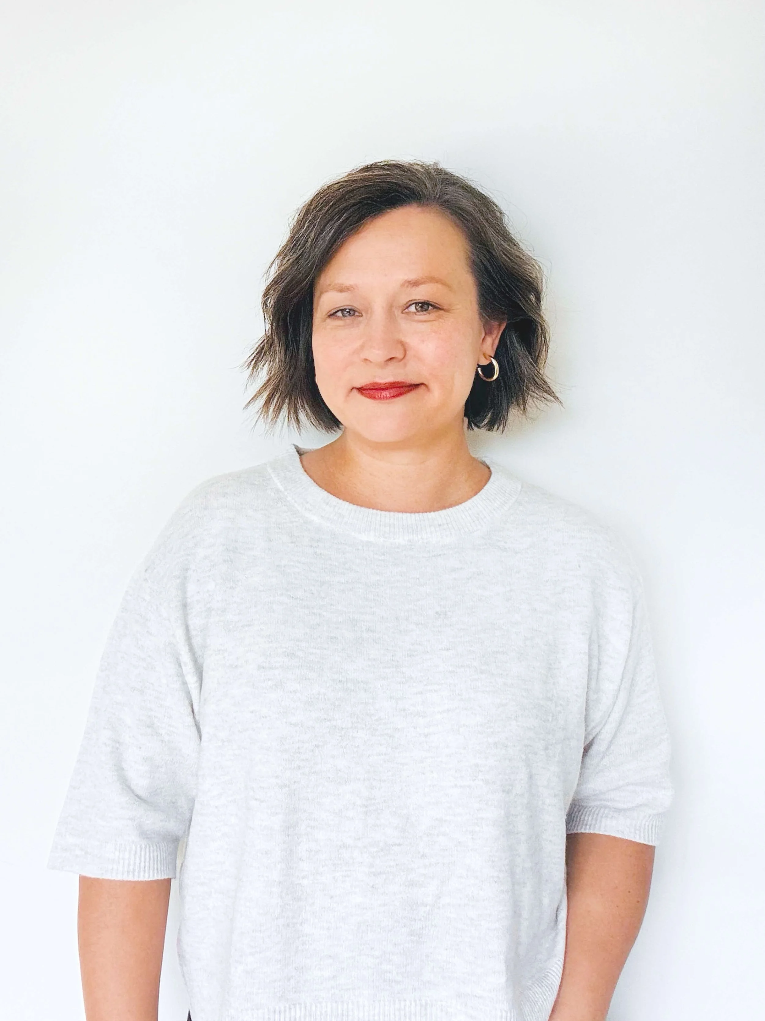 A woman with short, wavy brown hair and hoop earrings wearing a light gray sweater, standing against a plain white wall.