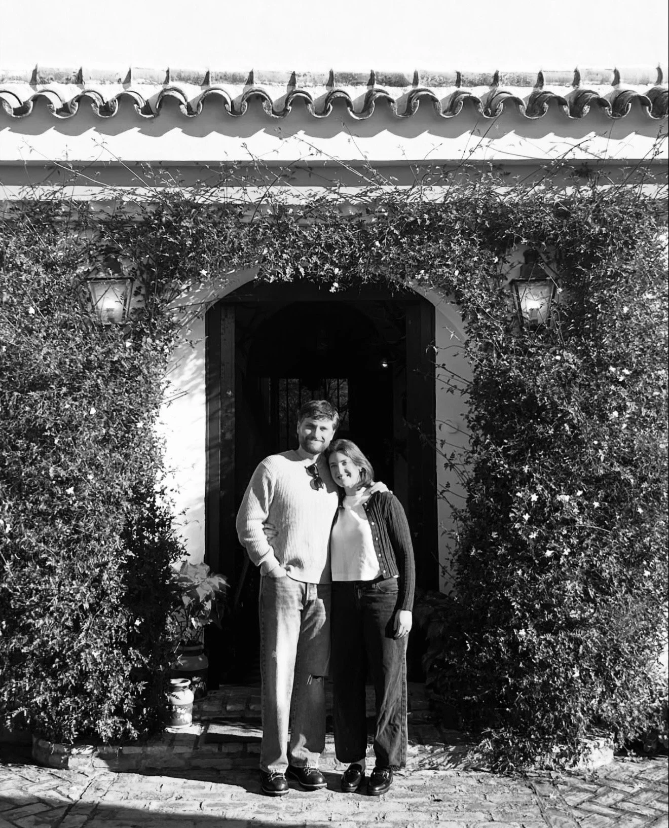 A black and white photo of a Drew and Sav standing close together under a spanish villa doorway decorated with bushes and vines, with two lanterns hanging on each side of the arch.