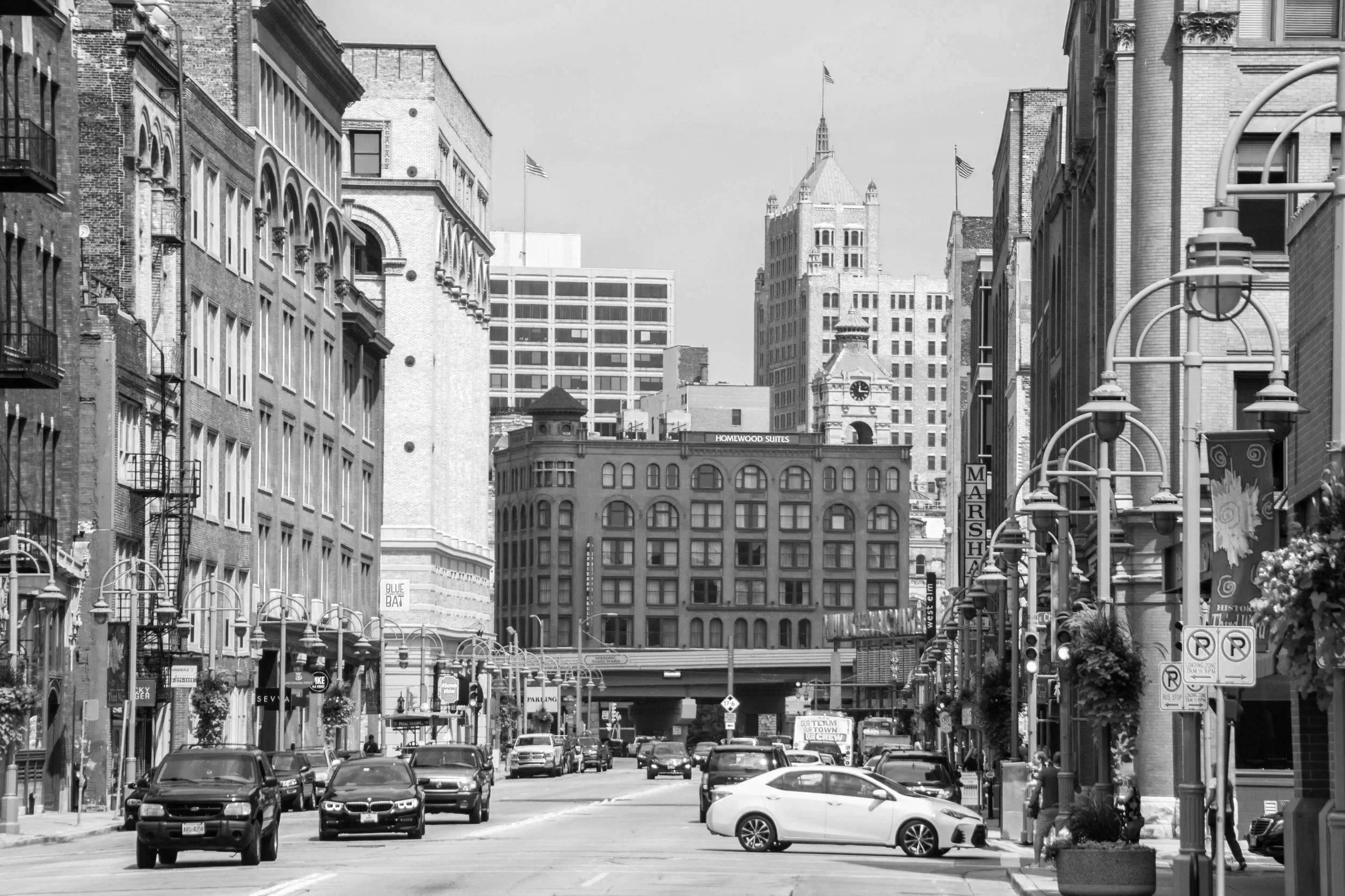 Black and white photo of a street in downtown Milwaukee's Historic Third Ward with cars parked and driving, buildings, street lamps, and pedestrians.
