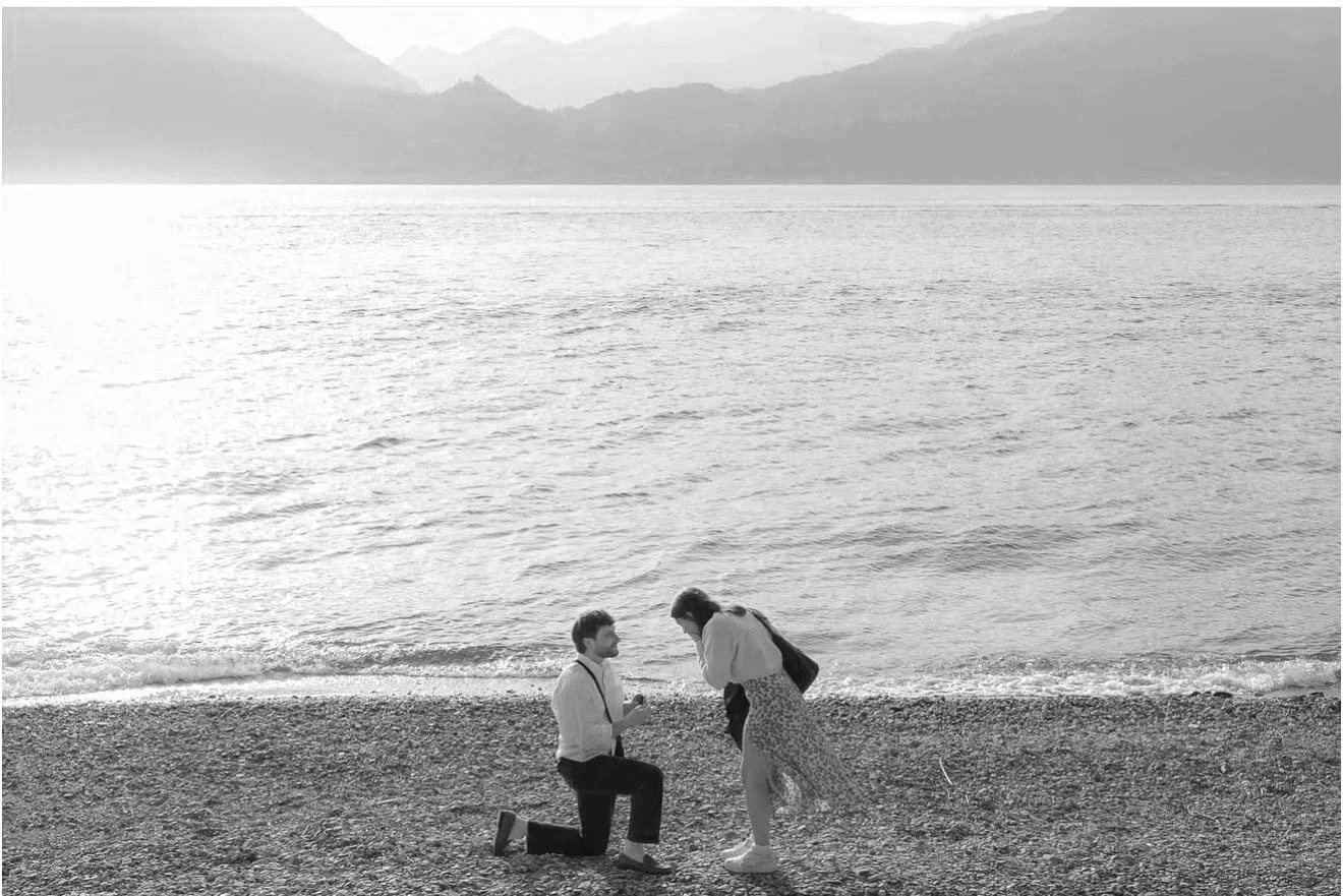 Drew proposing to a Sav on a beach in Italy, with mountains in the background.